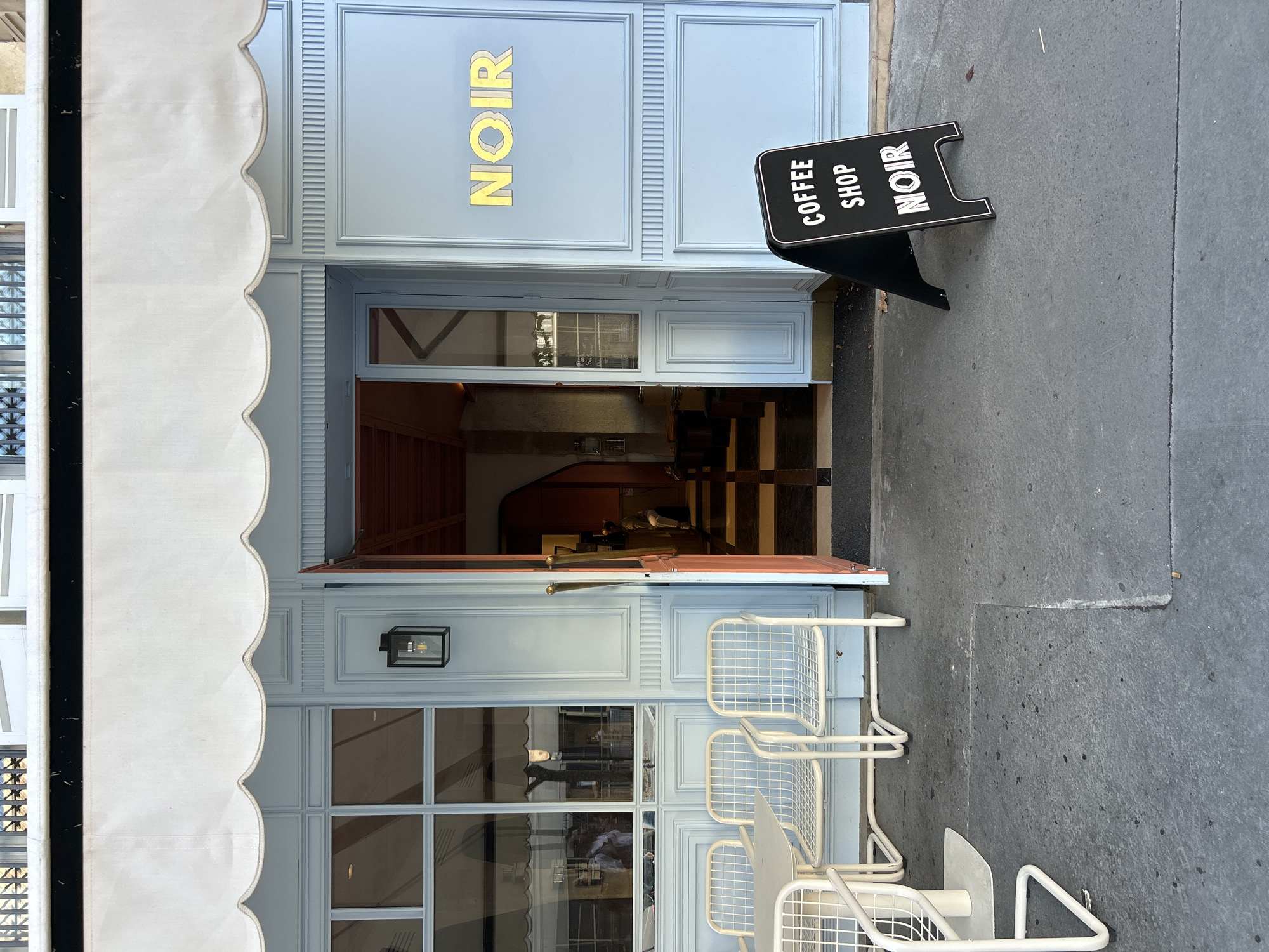 The image depicts the exterior of a cafe named "NOIR" in Paris, France. The cafe has a light blue facade with decorative molding and large windows. A white awning with a scalloped edge extends over the entrance. Outside the cafe, there are several white metal tables and chairs arranged on the sidewalk. A black A-frame sign in front of the cafe reads "COFFEE SHOP NOIR". The entrance door is partially open, revealing the interior of the cafe, which has a checkered floor and a dimly lit ambiance. A person is visible inside the cafe, sitting at a counter. The sidewalk is made of concrete and shows signs of wear, with some scattered debris. The overall impression is of a stylish and inviting Parisian cafe.