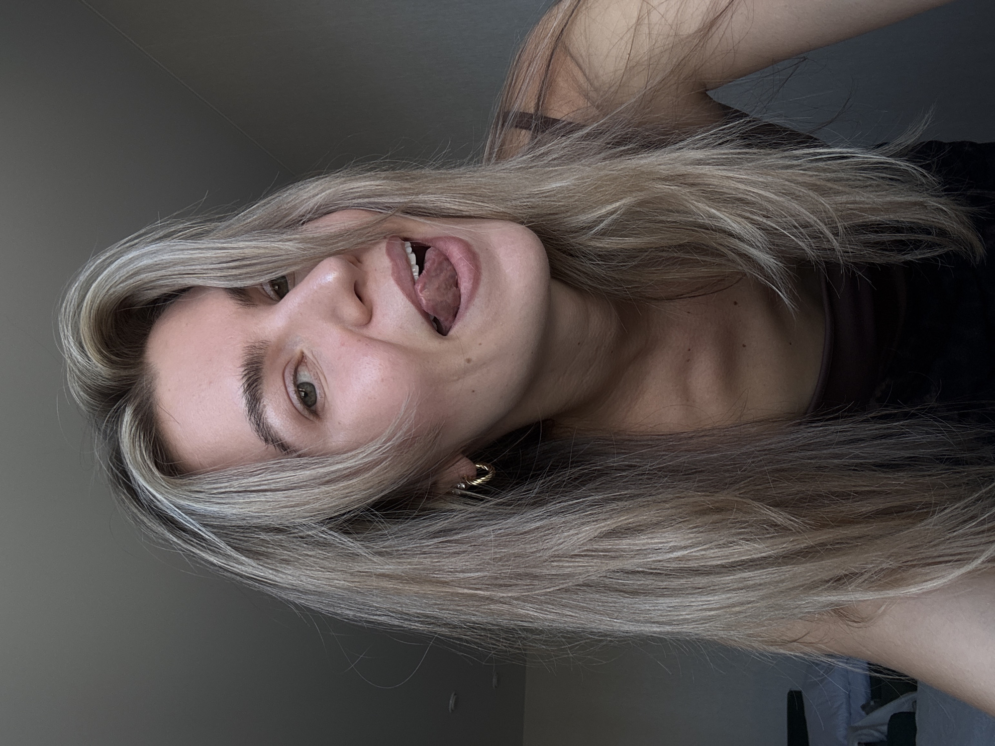 A woman with long blonde hair is seen from a low angle, lying down with her head tilted towards the camera. She has one eye visible and her mouth is open with her tongue extended. She is wearing an earring. The background consists of a plain gray wall.