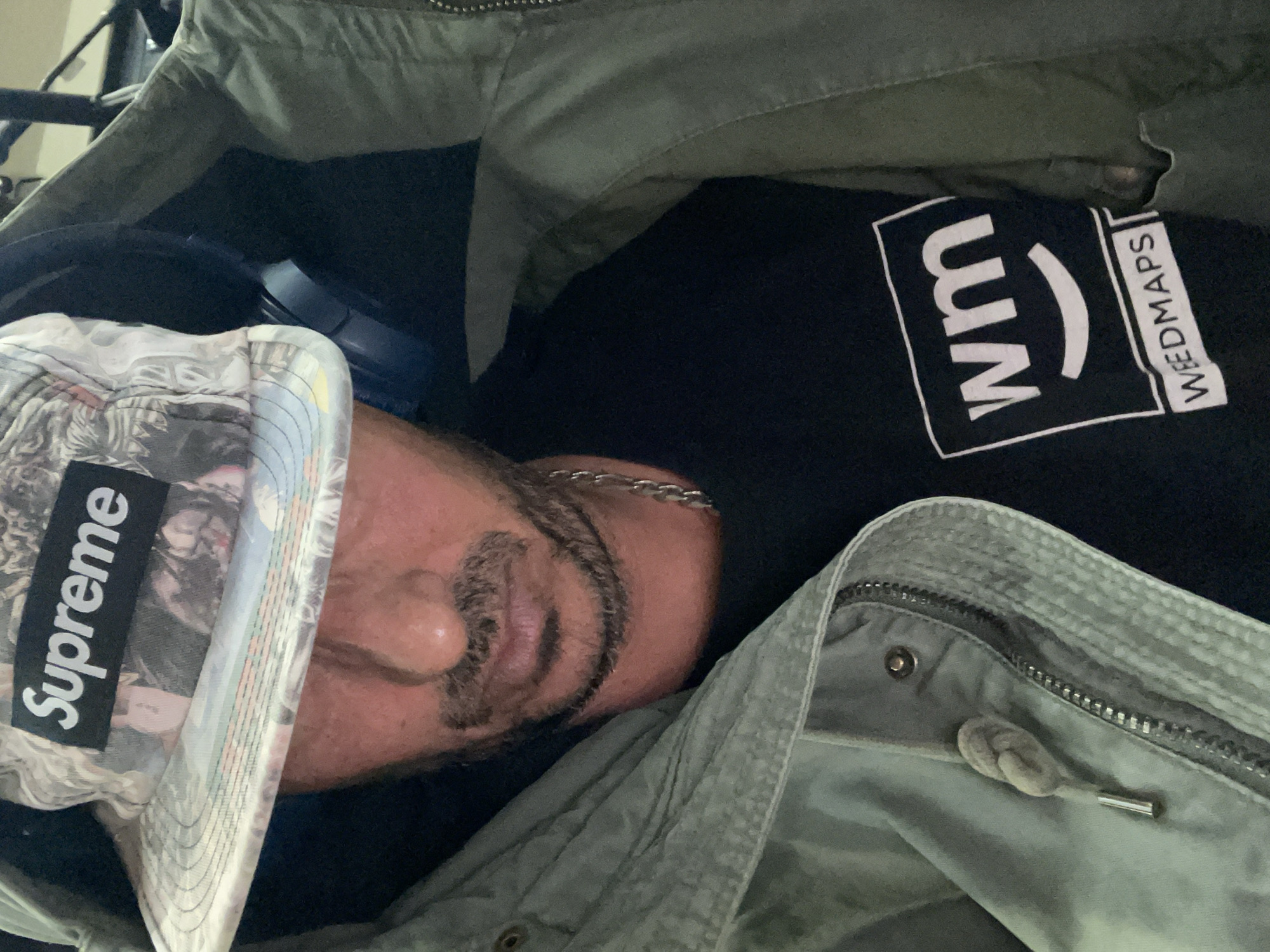 A medium-shot, close-up image from a slightly overhead perspective shows a person reclined, possibly sleeping, while wearing various articles of clothing and accessories. The person is wearing a Supreme-branded hat with a busy print, casting a shadow across their face, particularly the eyes. They also wear a black t-shirt with a white "WM" logo in a square with "WEEDMAPS" printed in smaller letters to the right. A silver chain is around their neck. Over the person, there is a light-colored, possibly faded green jacket with a zipper. Blue over-ear headphones are situated in the background to the left.
The context of the location in Barcelona, Spain cannot be determined from visual elements within the image.
