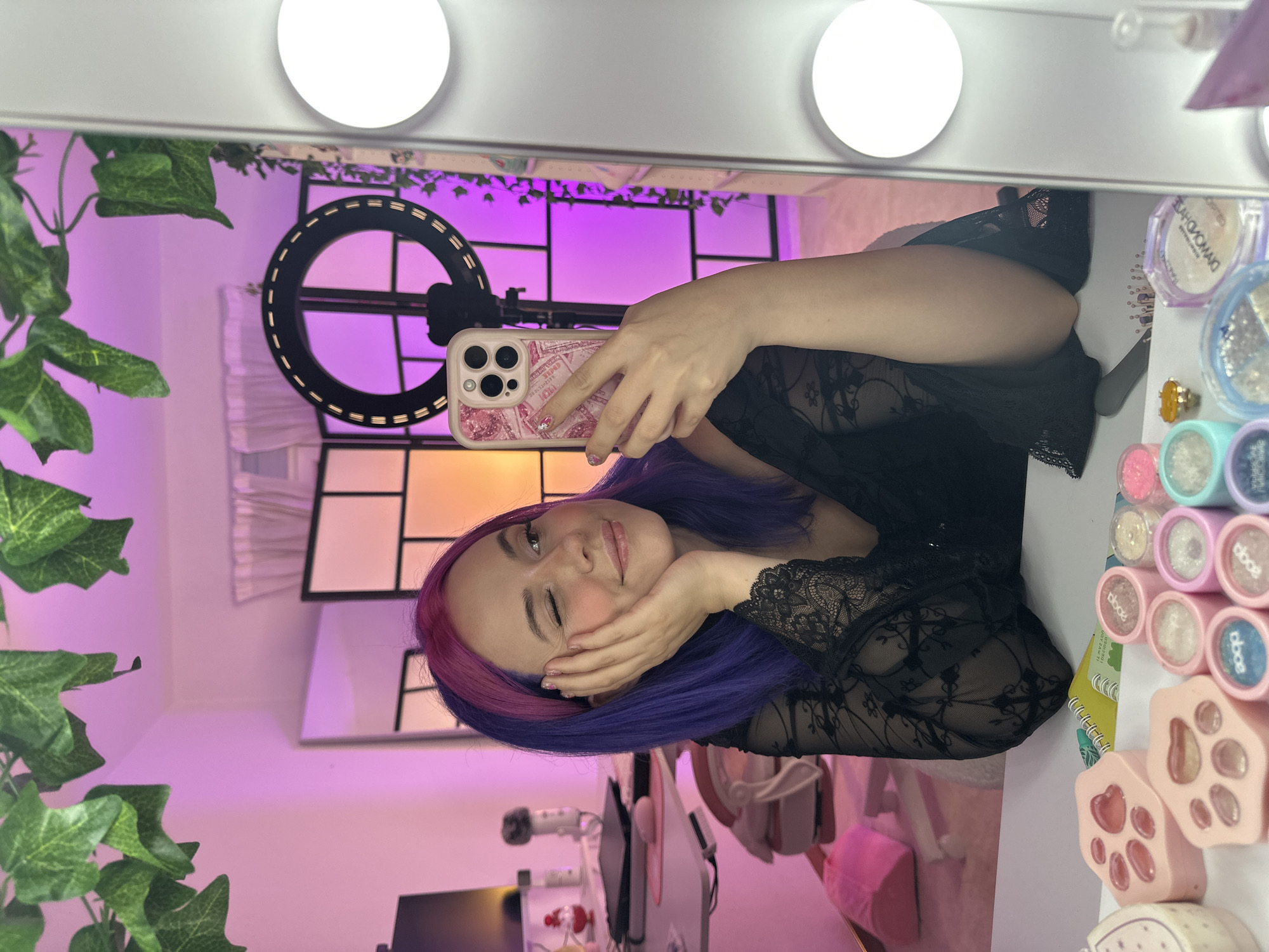 A young woman with purple and pink dyed hair is taking a selfie in a mirror. She is wearing a black lace top and is leaning her cheek on her hand. Her eyes are closed and she is smiling. In the background, there is a ring light, a folding screen, and various items on a desk, including makeup and a phone. The room is lit with pink and purple lights. The image is a selfie taken in a bedroom or vanity area, likely in Oviedo, Spain.