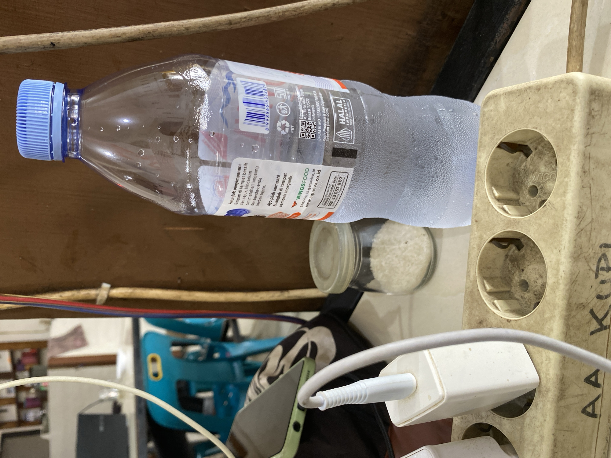 The image shows a cluttered indoor scene. A half-empty, condensation-covered plastic water bottle with a blue cap lies horizontally. Behind the bottle is a small jar containing rice. An extension cord is visible to the right, with a white phone charger plugged into one of the sockets. Cables and charging cords are present near a smartphone, suggesting electronic devices being charged. A blue plastic chair can be partially seen in the background. The setting appears to be a room with items stored under a table. Based on the bottle labeling, the bottle and thus the image is probably from Indonesia, but there is nothing indicating the location in the city of Lhokseumawe.