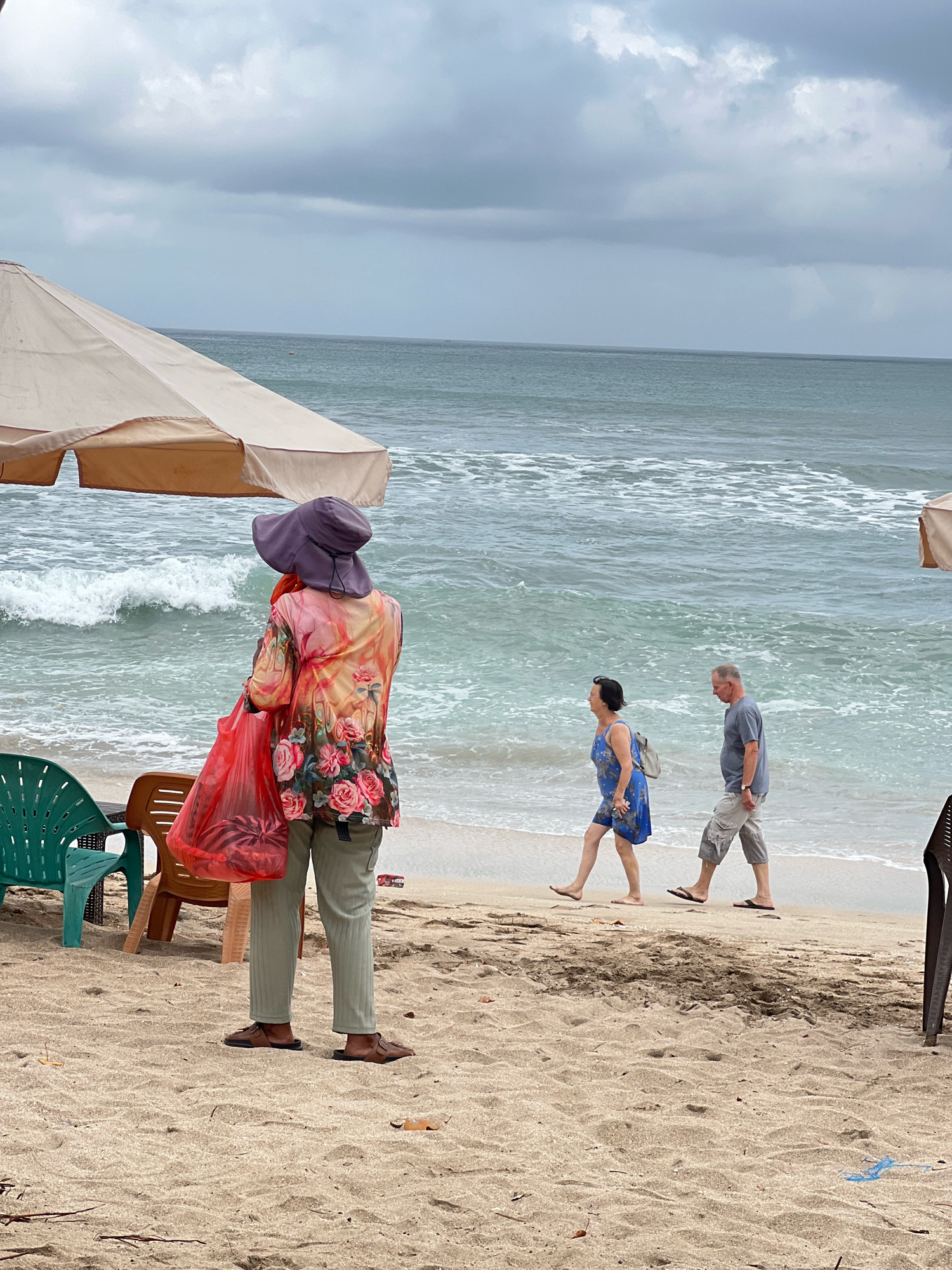 The image shows a cloudy day on Kuta Beach, Indonesia. In the foreground, a person wearing a wide-brimmed hat, a floral-patterned shirt, and light-colored pants stands with their back to the camera, holding a bright red plastic bag. They are wearing brown sandals. To their left, a green plastic chair and a brown plastic chair are visible. In the middle ground, a couple walks along the shoreline. The woman wears a blue floral dress and carries a small backpack, while the man wears a gray t-shirt and light-colored cargo shorts. The ocean is calm with gentle waves, and the sky is overcast with gray clouds. The beach is sandy, with scattered debris.