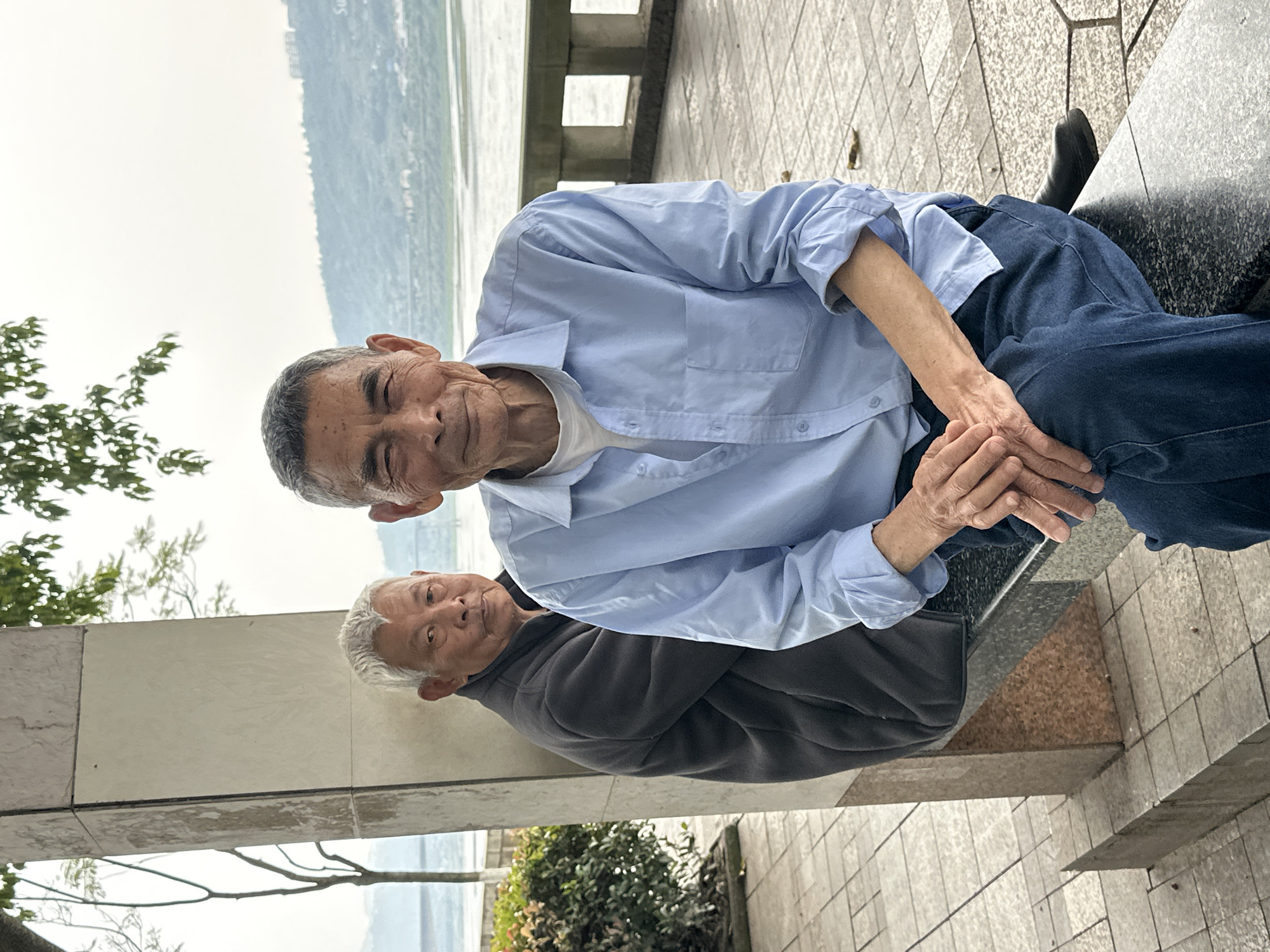 Two elderly Asian men are sitting outside in Jiangjin, China. One man is sitting on a ledge in a light blue button-down shirt and dark blue jeans. He is holding his hands together and looking at the camera. The other man is standing behind him and to his left, leaning against a gray concrete pillar. He is wearing a gray jacket and has white hair. In the background, there is a body of water and some green trees and bushes. The ground is paved with light gray tiles.