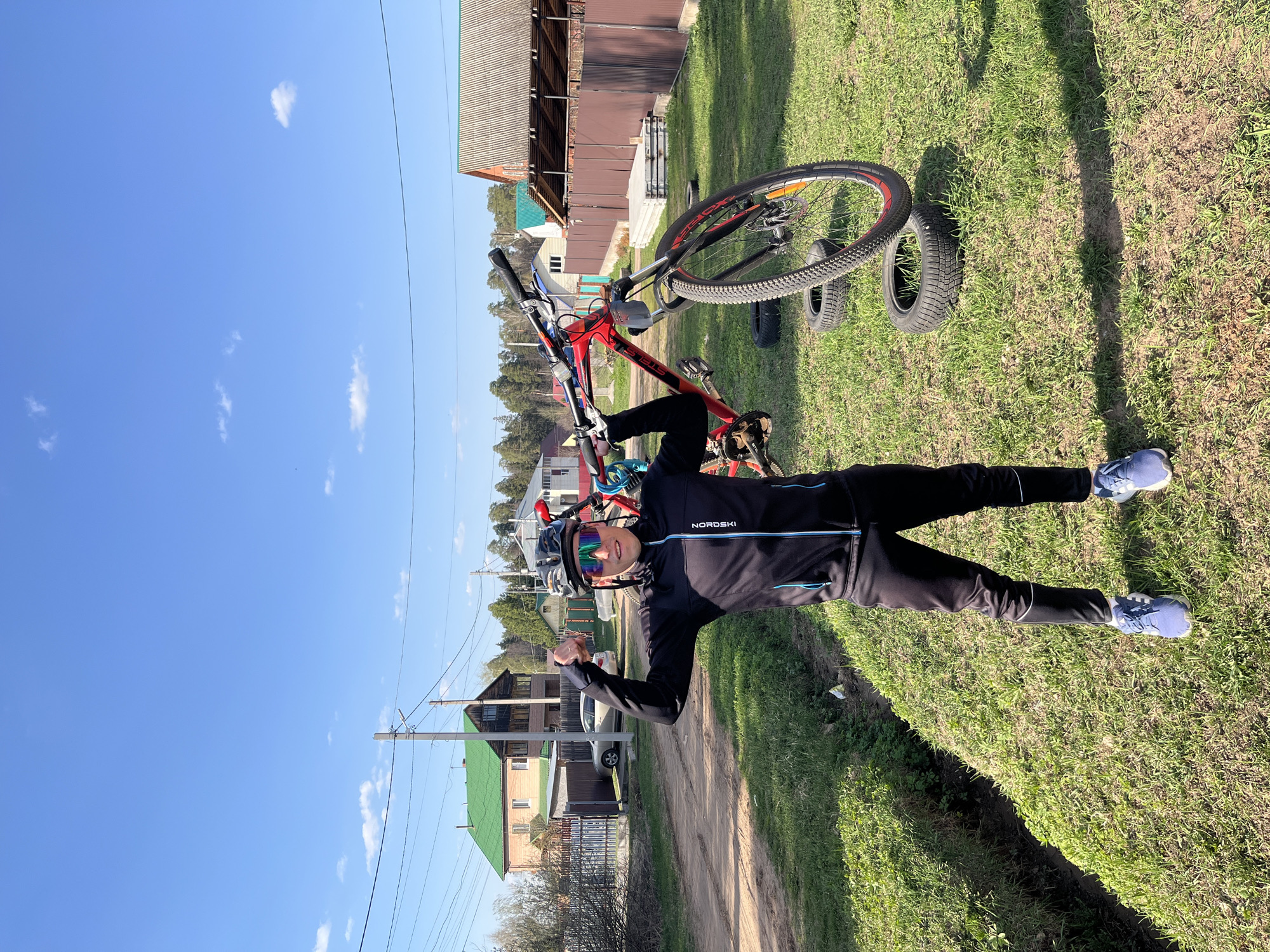A man wearing a black cycling outfit and helmet is holding a red bicycle above his head in a rural area, posing for a photograph. The man is standing on a grassy area adjacent to a dirt path that runs alongside a residential area with houses. The sky is clear and blue with some scattered clouds. Some houses have a green roof. Based on the rural atmosphere and general layout of buildings, it is likely the location is in a suburb of the city of Chaikovsky, Russia. Additional tires are present near the bicycle on the grass.