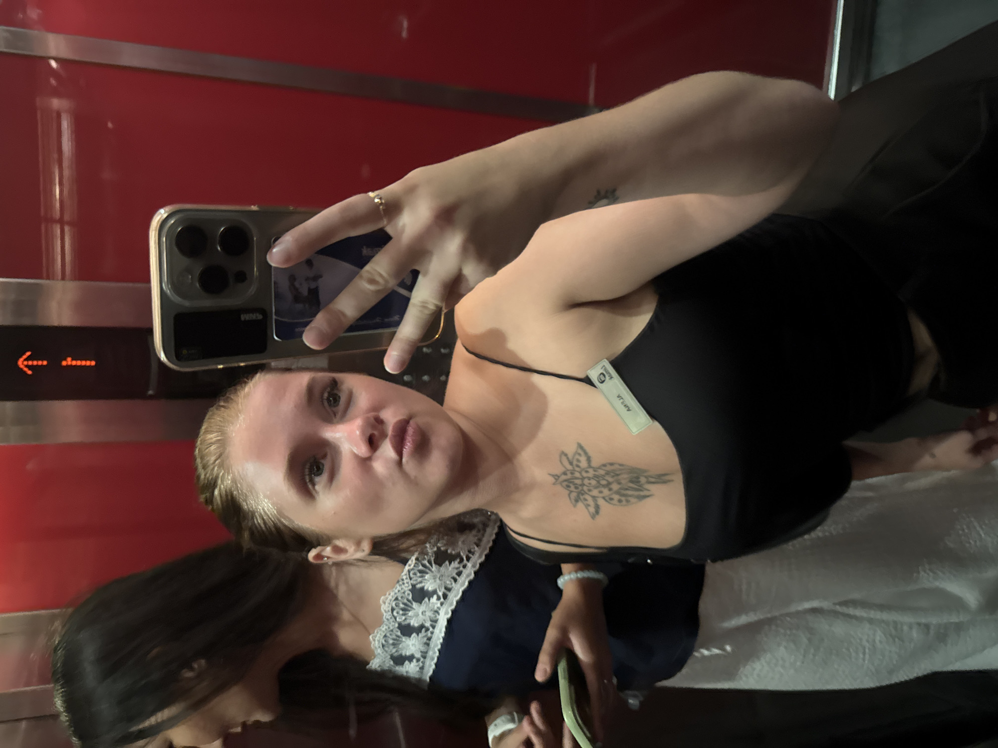 Two individuals are inside an elevator with red walls. The person in the foreground, a female with blonde hair tied back, is taking a selfie with her smartphone. She is wearing a black tank top and has a tattoo of a flower on her chest. A name tag that reads "AMELIA" is pinned to her top. She is making a "duck face" pose and holding up three fingers on her right hand. The person in the background, also female with dark hair, is partially visible, wearing a dark blue top with white lace trim. She is holding a green object, possibly a phone or wallet, in her left hand and wearing a white bracelet. The elevator doors are closed. The scene is set in an urban environment, specifically an elevator in Aksu, Turkey.