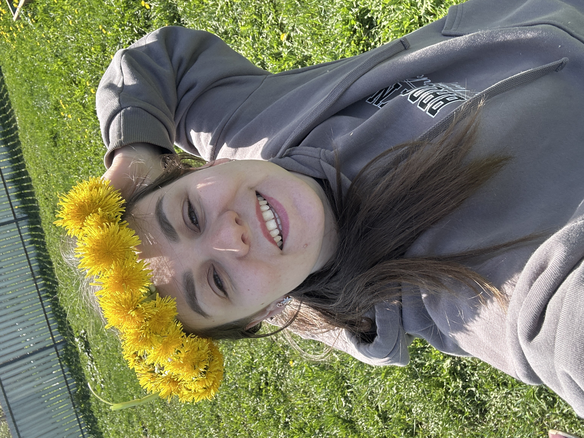 A young woman is lying on her back in a grassy area, possibly in a park. She is wearing a gray hoodie with white lettering on the front. She is also wearing a wreath made of yellow dandelions on her head. She is smiling and looking directly at the camera. The location is likely somewhere in the city of Chaikovsky, Russia, as that information was provided in the prompt. The sun is shining, creating shadows on the grass and the woman's face and clothing. In the background, there is a metal fence.