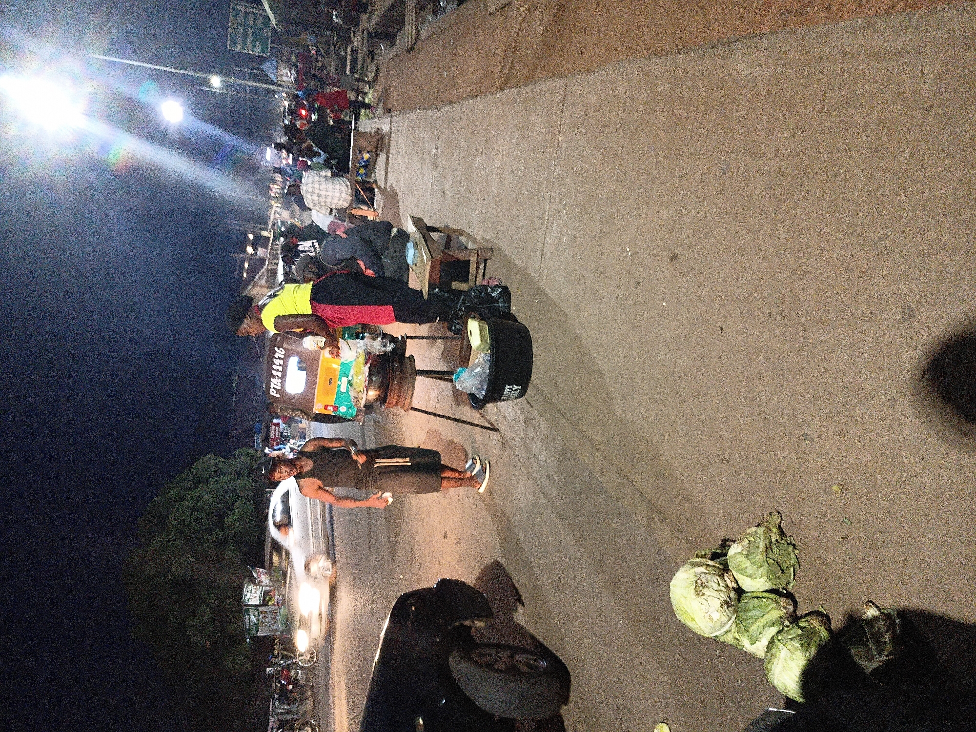 A nighttime street scene in Bukuru, Nigeria, shows a busy roadside with vendors and pedestrians. In the foreground, a man in a dark tank top and shorts stands near a food cart, holding a small white object. To his left, several heads of lettuce are piled on the asphalt. Behind him, a woman in a yellow shirt and red skirt stands by the food cart, which has a steaming pot. A yellow and green tricycle is parked beside the cart. In the background, a white car with illuminated headlights drives by, and numerous people are gathered, some sitting on benches. The street is lit by bright overhead lights, casting long shadows and creating a vibrant atmosphere.