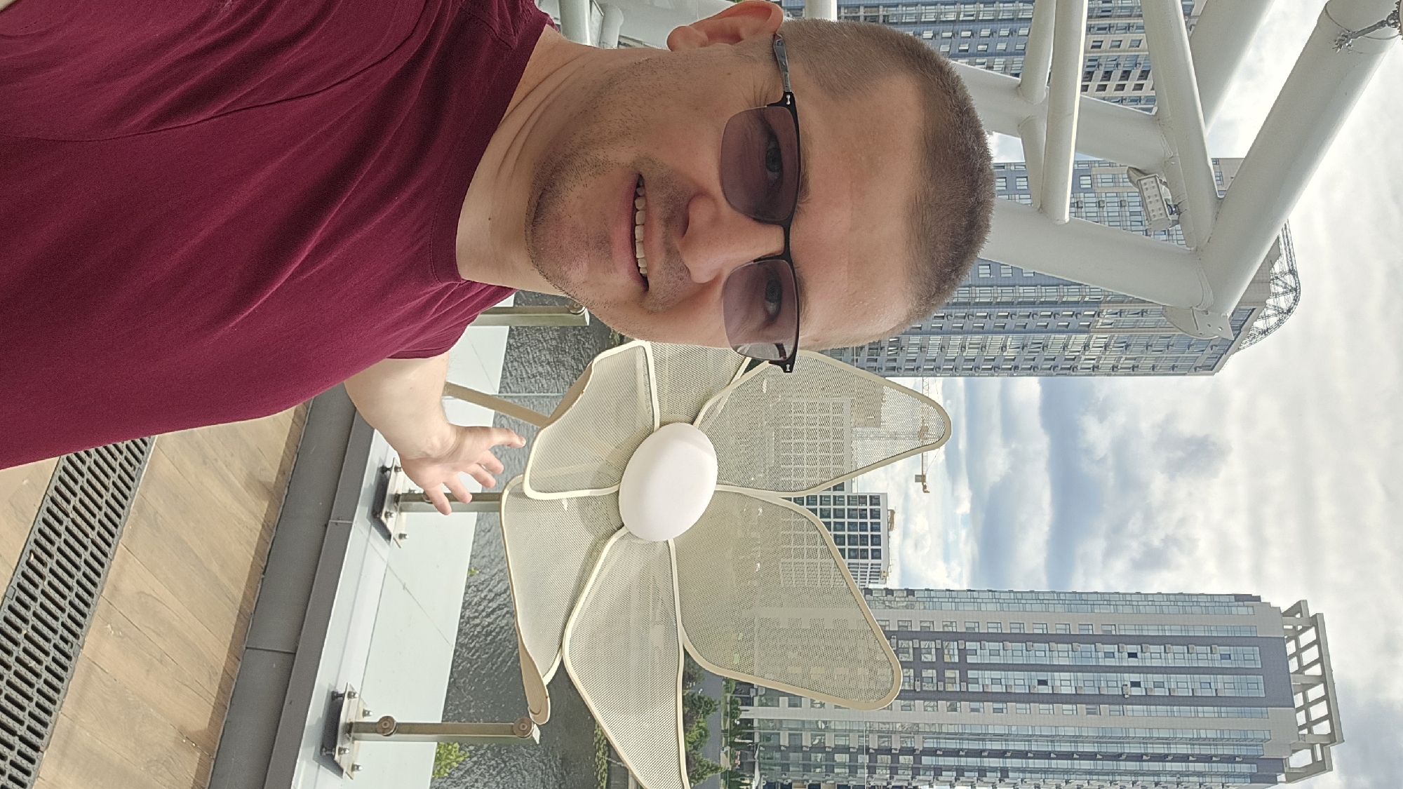 In the foreground, a man with short hair and glasses smiles at the camera. He is wearing a maroon t-shirt and his left arm is extended outwards, with his hand open. To his right, a large, decorative flower sculpture made of metal mesh with a white oval center is visible. Behind the man and sculpture, a body of water is present, with a walkway and railing along the edge. In the background, two tall, modern buildings with numerous windows dominate the cityscape. The sky is cloudy. The scene is likely a public outdoor space or a rooftop terrace in Kyiv, Ukraine, possibly near a river or waterfront area, given the architectural style and the presence of the water.