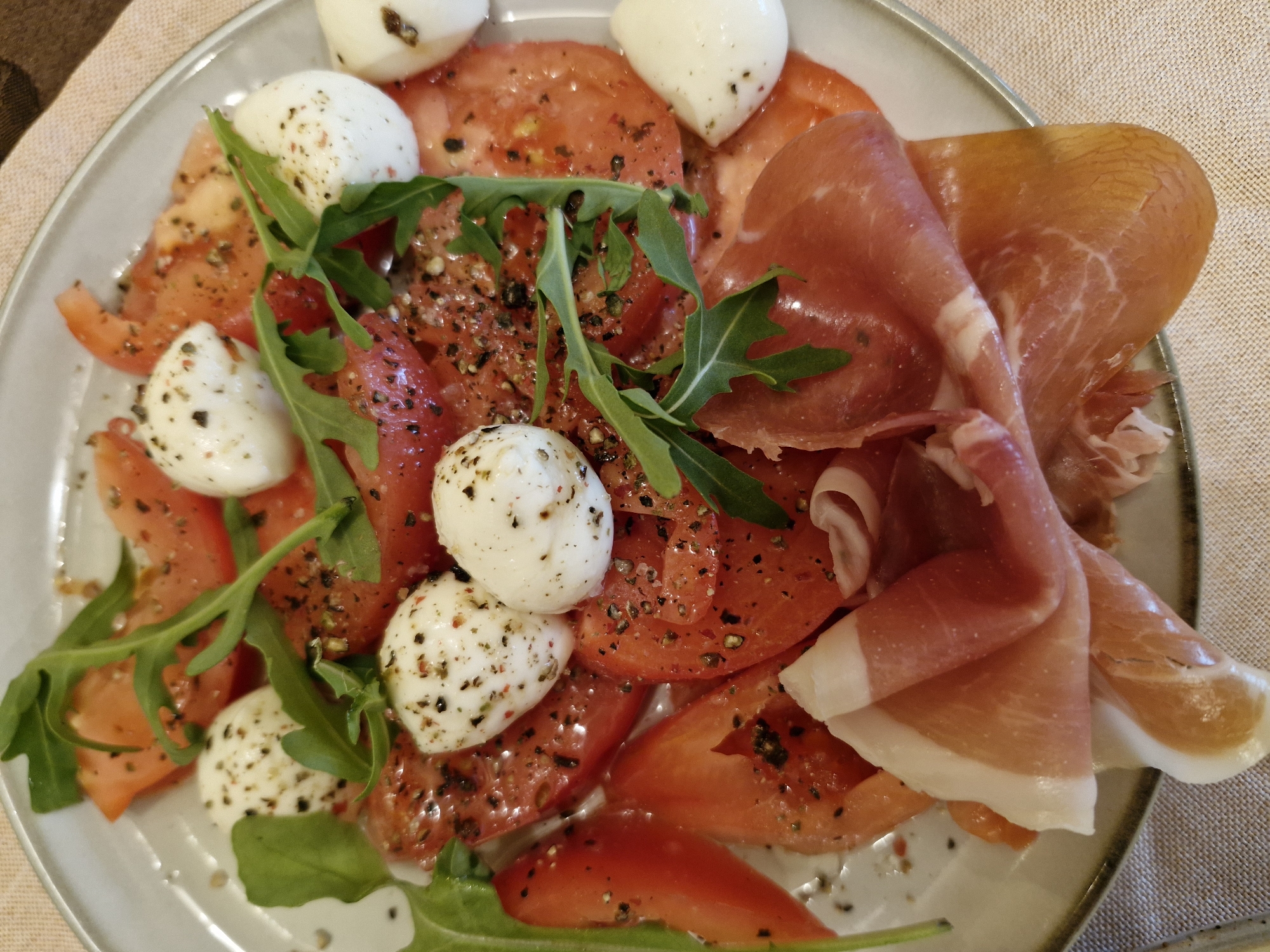 An overhead, close-up image displays a prepared Italian-style dish on a round, shallow, light gray plate. The plate contains a base of numerous red tomato slices of varying shapes, some wedge-cut and others rounded. Scattered across the tomatoes are approximately ten small, white, spherical mozzarella balls, each coated with dark and reddish-brown speckled seasoning, likely ground pepper and dried herbs. Dark green arugula leaves are interspersed among the tomatoes and mozzarella. At the bottom right and center-bottom of the plate, several thin, folded slices of cured ham (prosciutto) with pink, red, and translucent white fatty areas are arranged. The plate rests on a light beige, tightly woven fabric surface, exhibiting a distinct grid pattern. No other environmental details are visible. There are no people present, and no specific actions are depicted. The location in the city of Unknown, Unknown, cannot be determined from the image.