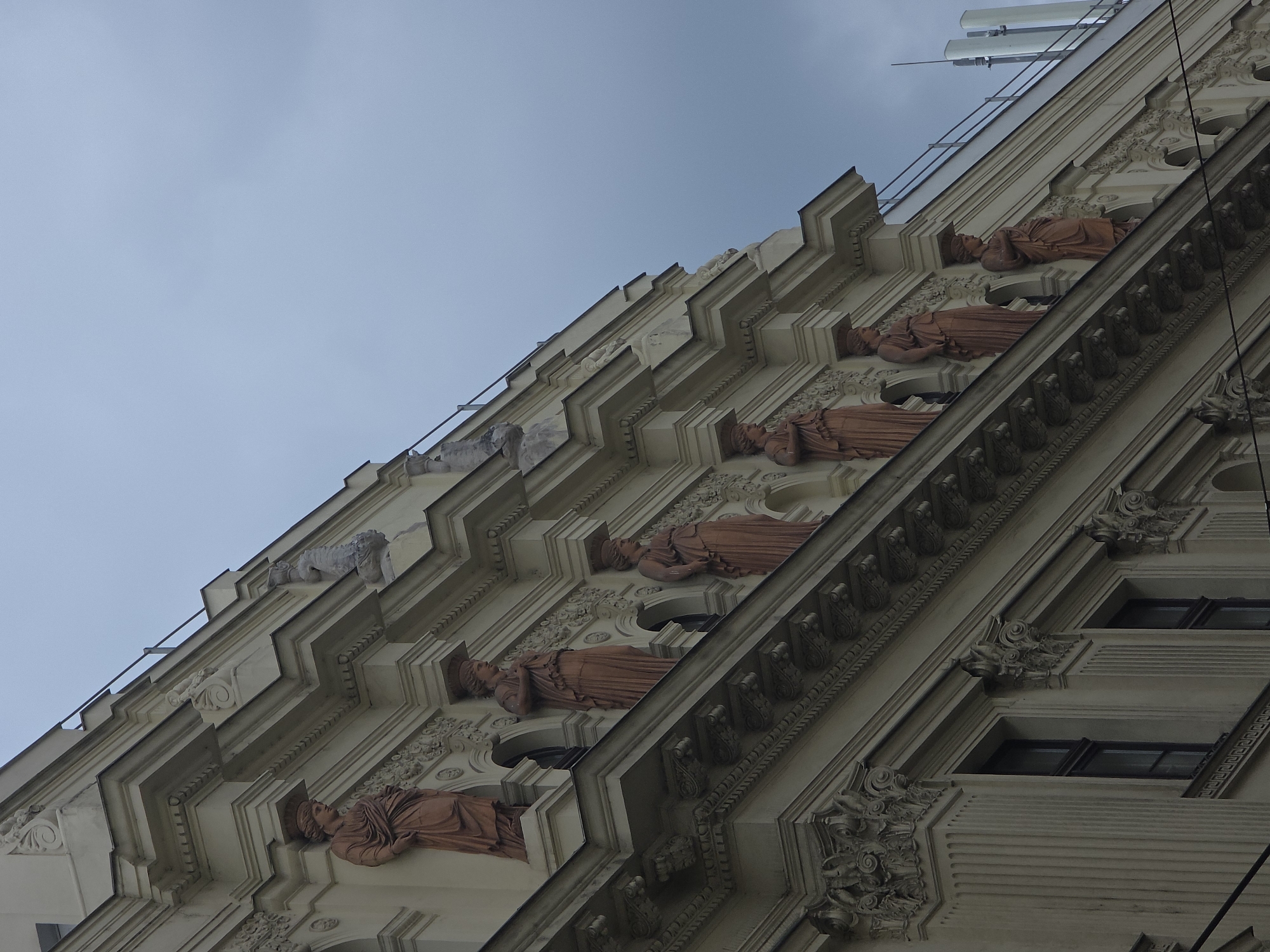 A low-angle shot captures the ornate facade of a cream-colored building, its upper levels adorned with classical statues. The statues, depicted in a terracotta hue, are of women draped in flowing robes, each standing within a recessed alcove. They are positioned along the top edge of the building, beneath a projecting cornice. The architectural style suggests a historical European city, with intricate carvings and decorative elements accentuating the building's exterior. The sky above is overcast and a muted gray, with hints of a dark, moody atmosphere. Modern telecommunication equipment is visible in the upper right corner of the image, contrasting with the classical architecture. The scene is devoid of people or any visible activity.