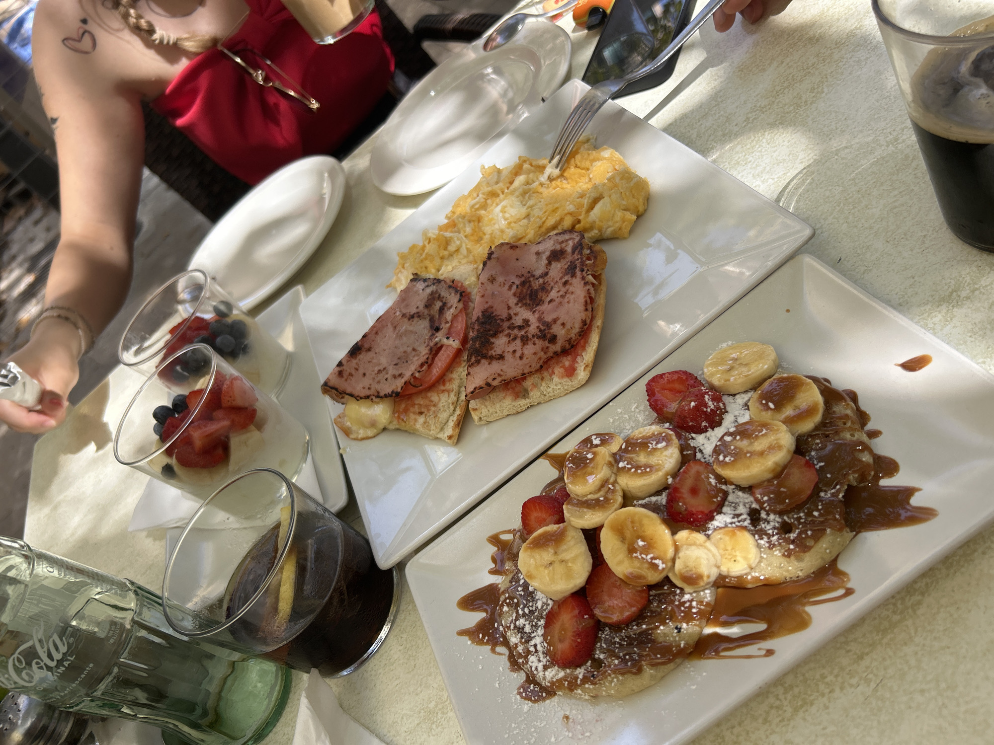 The image depicts a table at an outdoor cafe, likely in Manacor, Spain, as indicated by the presence of a Coca-Cola bottle with Spanish text and a general Mediterranean ambiance. On the table are two white rectangular plates of food. The plate in the foreground contains a waffle or pancake topped with sliced strawberries, bananas, and drizzled with caramel sauce and powdered sugar. To the right, another plate holds scrambled eggs and a sandwich with ham and tomato slices on toasted bread. Two glasses of beverages are visible: one filled with a dark liquid, possibly cola with a lemon slice, and another containing a portion of yogurt with berries. In the background, the arm and shoulder of a woman wearing a red top are visible, along with a partial view of another person. The setting appears to be a bright, sunny day.
