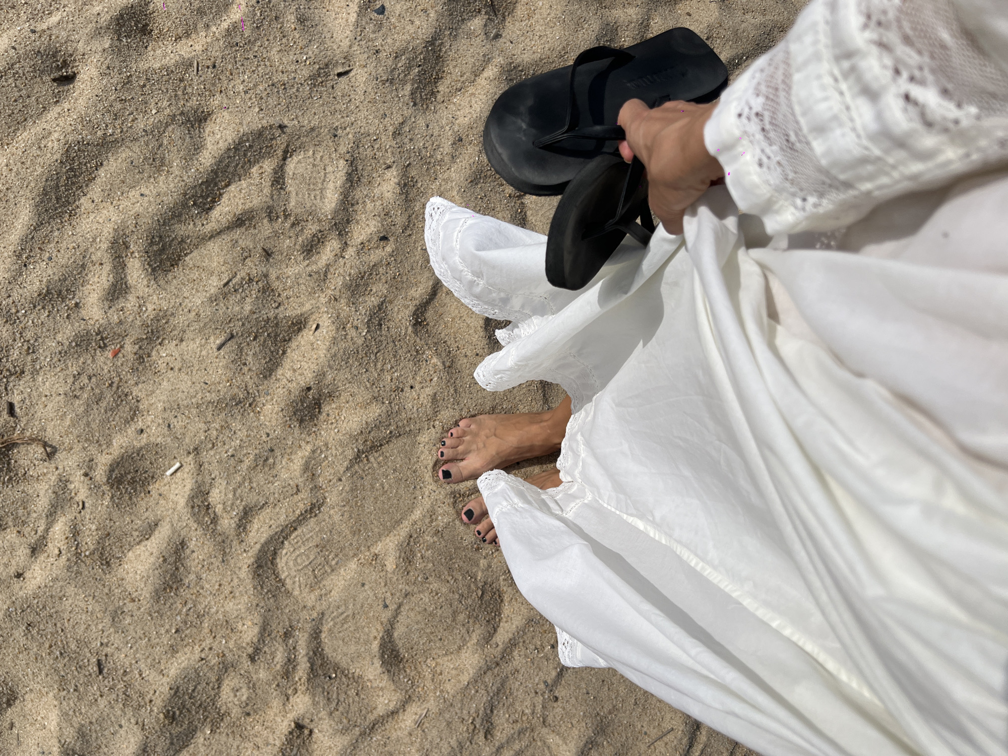 This image presents an overhead perspective of a person's feet and lower attire on a sandy surface in Nha Trang Ward, Vietnam. In the mid-left of the frame, a pair of bare feet with dark-painted toenails are positioned on light brown sand. A white, flowing garment, likely a dress or skirt, is draped around the feet, with its fabric being held up by a right hand in the lower-right quadrant. The garment features white lace detailing on the sleeve near the wrist. The hand also holds a pair of black thong-style flip-flops, with the brand name "HAVAIANAS" partially visible on one strap. The surrounding sand exhibits textured ripples and contains various small debris, including a white cigarette butt in the upper-middle, several dark small sticks, and minute colored plastic fragments. The scene is illuminated by bright, natural daylight.