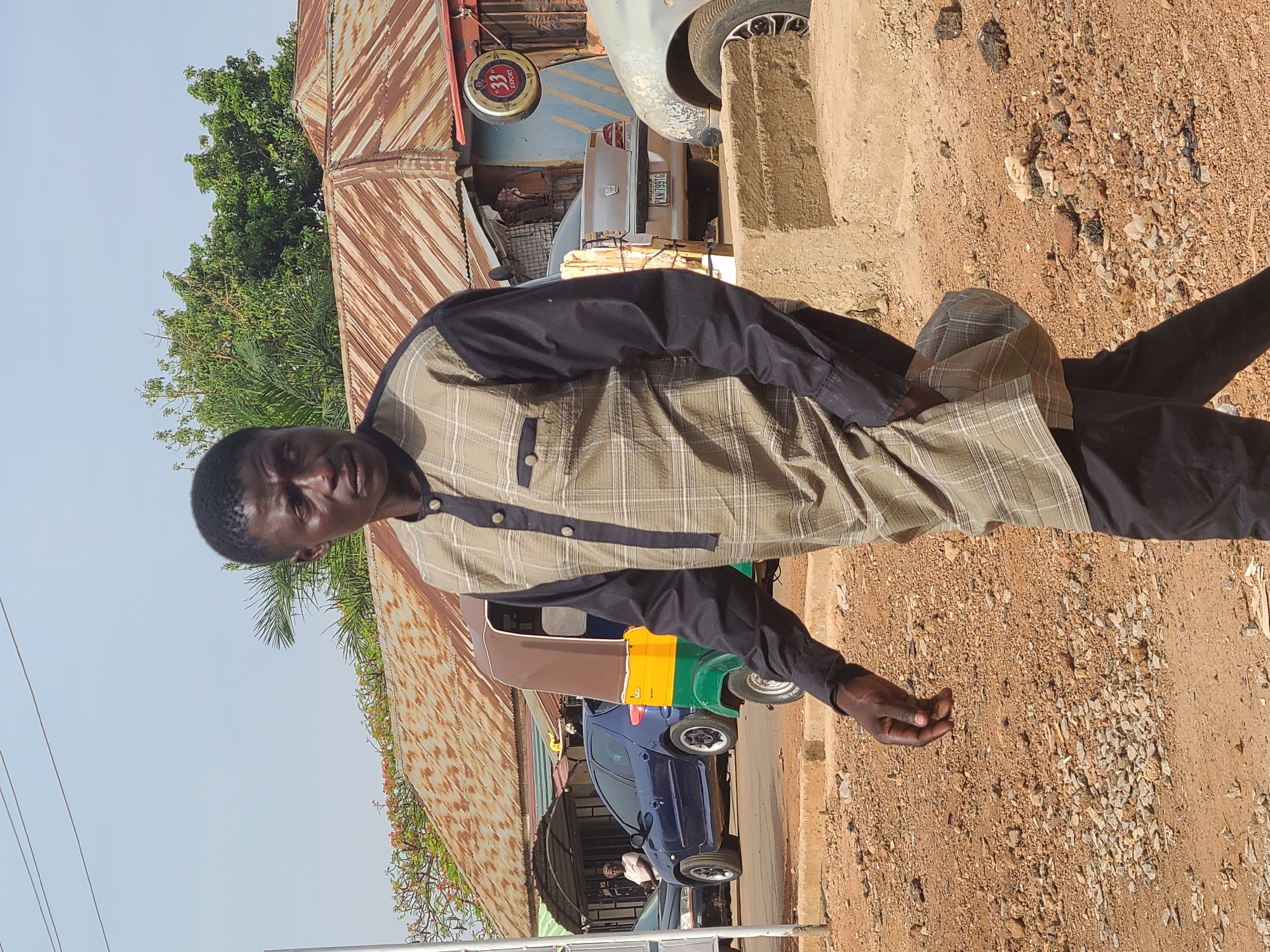 A man stands on a dirt road in Jos, Nigeria. He wears a black shirt with a beige and black plaid overshirt. He is looking directly at the camera. Behind him is a street with a parked blue car, a yellow and green vehicle, and a silver car. A building with a rusty corrugated metal roof and a "33 Export" sign is visible, as well as green trees.