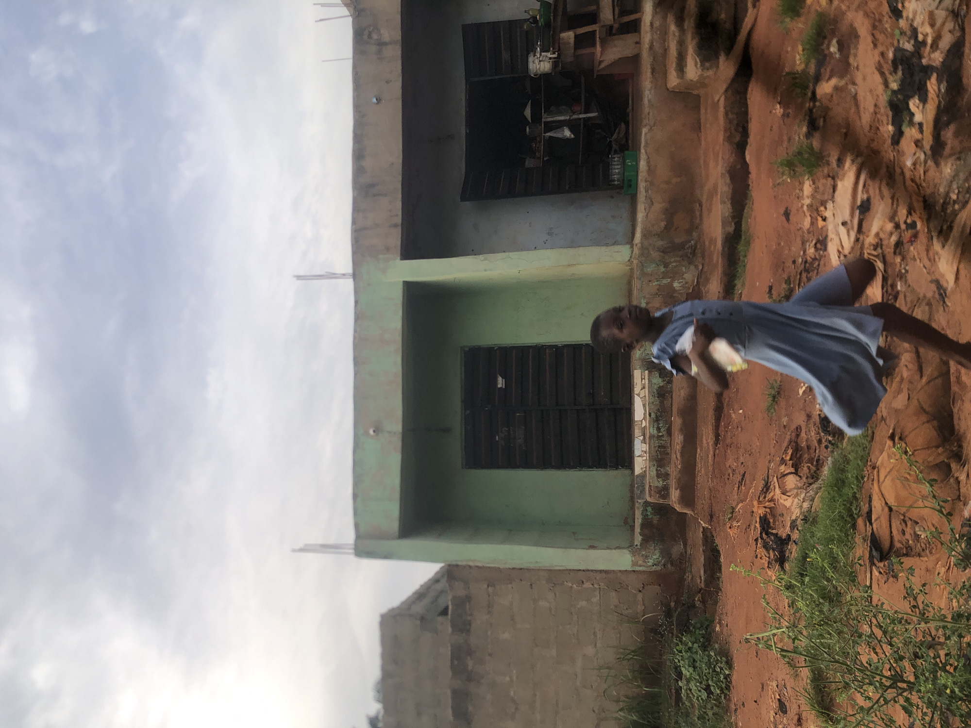 The image captures an outdoor scene, likely in Ukwu-Abwa, Nigeria, under an overcast sky. In the foreground on the right, a young person, possibly a girl, stands wearing a light blue dress. She holds a light-colored object in her hands and looks towards the camera with a neutral expression.
Behind her and occupying the mid-ground on the left, is a two-story building. The lower level is painted a light green, while the upper level appears to be a faded grey or brown. Both levels feature dark, shuttered windows. A partially constructed wall made of concrete blocks is visible in the lower-left corner of the frame.
The ground beneath and around the subjects is composed of reddish-brown dirt, characteristic of laterite soil, with scattered patches of green vegetation and some debris. The cloudy sky fills the upper portion of the frame, indicating daytime. There is no visible text in the image.