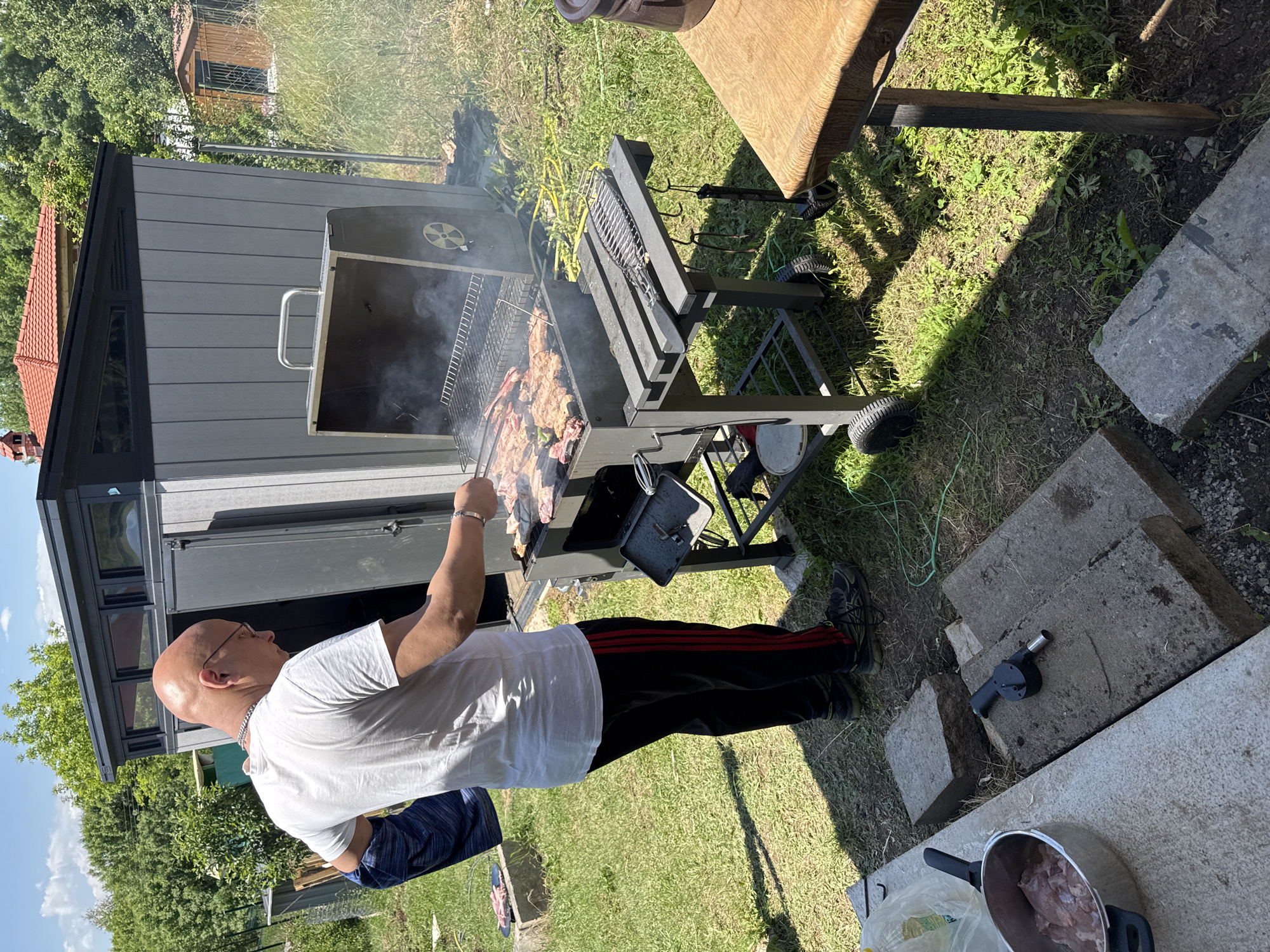 An outdoor scene in Dolna Dikanya, Bulgaria, depicts a male individual tending to a barbecue grill on a sunny day. The man, bald and wearing glasses, a white t-shirt, and black track pants with red side stripes, is positioned to the left of a dark grey charcoal grill, with smoke rising from it. He wears a silver-colored chain necklace and a silver-colored bracelet on his left wrist, and has a dark blue fabric draped over his left forearm. His right hand is near the cooking meat on the grill.
The grill, equipped with side shelves, holds multiple pieces of meat cooking over coals. A metal fish-shaped grilling basket and a black spatula are on the right side shelf. Immediately behind the man and the grill is a modern, dark grey, vertically paneled shed with small horizontal windows and a dark roof. One of its double doors is slightly ajar.
The ground is primarily covered in grass, with scattered patches of bare earth. In the foreground, concrete slabs serve as makeshift surfaces. A silver pot containing raw, light-colored meat (likely chicken) rests on one slab in the lower left, adjacent to a clear plastic bag. A black manual charcoal blower tool is situated on another concrete slab nearby. A green electrical cord or hose snakes across the grass in the midground. A partial view of a wooden table with a thick light brown top and dark legs is visible on the far right, holding a dark brown container.
In the background, lush green trees and bushes are prominent. Above the main shed, the red-tiled roof of a building is visible. To the right of the main shed, beyond a wire fence, a smaller, brown wooden shed or structure stands. The sky is blue with some scattered white clouds, and direct sunlight casts distinct shadows across the scene.