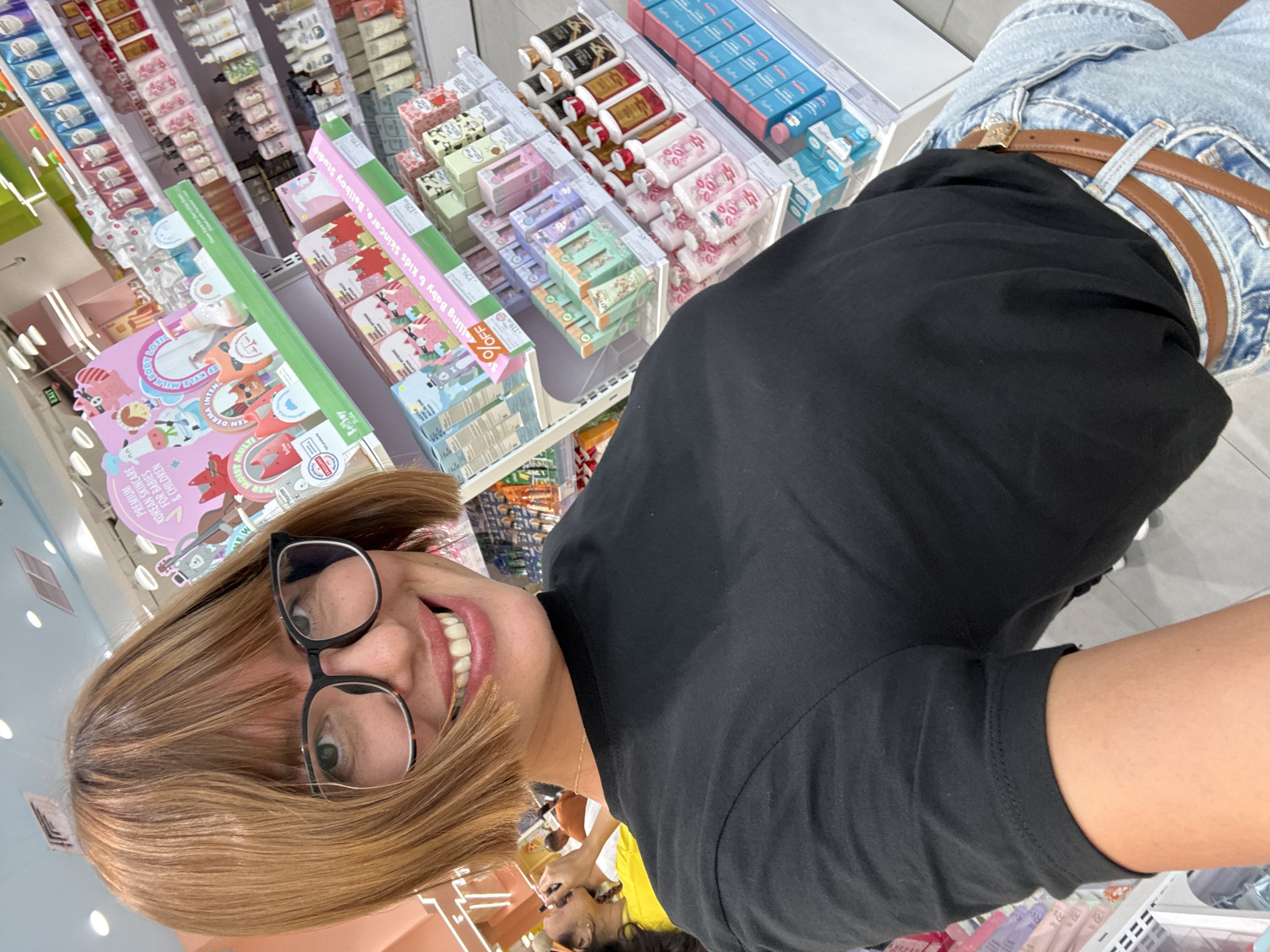 A selfie depicts a woman wearing glasses and a black shirt standing in a retail environment. In the background, shelves are stocked with various packaged goods, including what appears to be Korean skincare products for babies and children. The location is a store, specifically a cosmetics or skincare outlet, possibly in Kuta, Indonesia given the Korean skincare products, but this cannot be confirmed definitively from the image alone. The woman is smiling at the camera, and behind her, there's a glimpse of other people near a checkout counter, including a person in a yellow shirt. The image suggests an indoor shopping setting.
