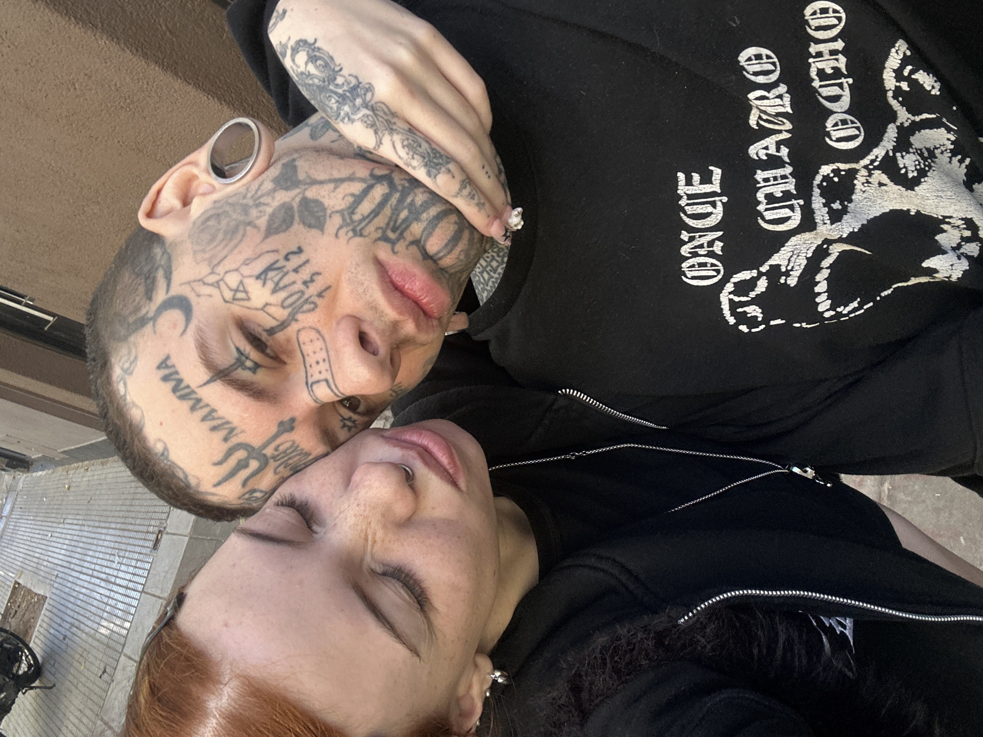 The image captures two individuals, a man and a woman, in a close and intimate embrace, taken from an overhead perspective during daytime in Buenos Aires, Argentina. The man is positioned to the left, his heavily tattooed face looking down towards the woman resting against him.
The man exhibits extensive facial, head, and arm tattoos, including script like "MAMMA" on his forehead, "TWO" on his cheek, and numbers "21" and "13," alongside various symbols and rose designs. He has large silver ear gauges, a septum piercing, and a small bandage is visible across his nose. He wears a black hooded sweatshirt featuring white text and a graphic. The visible text on his shirt clearly reads "ONCE CUATRO OCHO," which are Spanish for Eleven, Four, Eight. His hand, adorned with long, decorative fingernails, rests near the woman's face.
The woman, with reddish hair and visible freckles, has her eyes closed, conveying a sense of peace and intimacy. She wears a nose ring and a black zippered hoodie, with a silver chain visible around her neck.
The background suggests an outdoor setting with horizontal lines, possibly a paved ground or tiled surface. The overall impression is one of affection and connection between the couple, highlighted by their distinctive appearances.