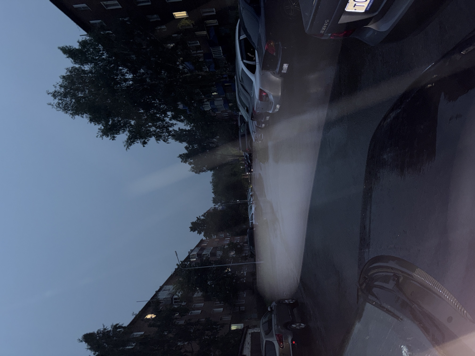 The image depicts a street scene in Chistopol, Russia, at dusk or night. The sky is a deep blue, indicating the transition from day to night, and the weather appears to be rainy, with water visible on the ground and reflecting light.
The primary subjects are several cars parked along a street lined with apartment buildings. The buildings are multi-story structures with many windows, some of which are illuminated, suggesting that people are inside. Trees with dark foliage are interspersed between the buildings and along the street, their leaves appearing wet from the rain.
The reflections on the wet pavement create streaks of light, possibly from streetlights or the headlights of vehicles. One car in the foreground has its windshield wipers visible, further confirming the rainy conditions. The overall mood is somber and tranquil, characteristic of a quiet evening in a residential area.
Visible text includes a license plate on one of the cars, which appears to have Russian characters and numbers. The context of Chistopol, Russia, is provided separately and is not directly inferred from visual cues within the image itself.