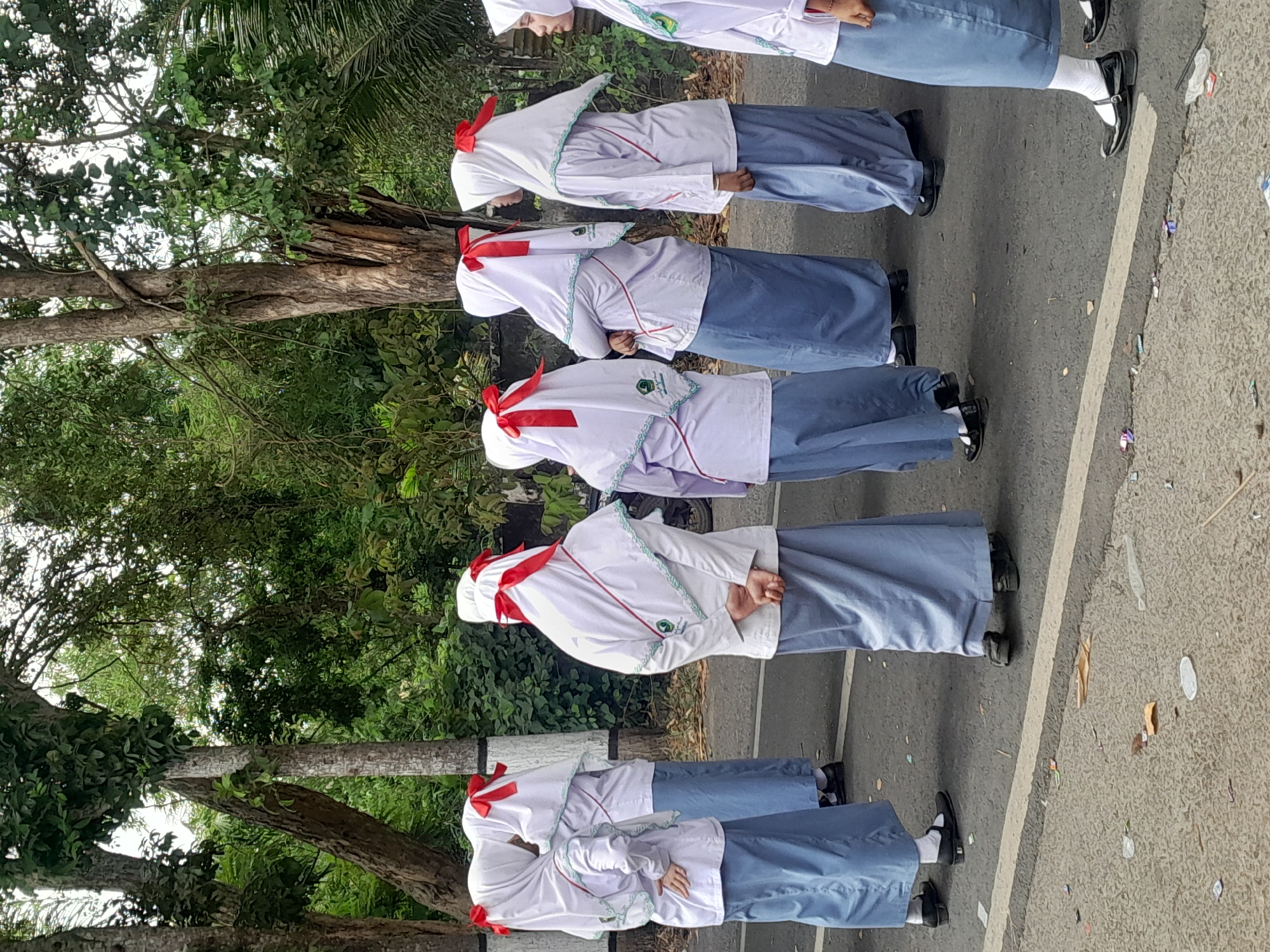 In Sempu, Indonesia, a group of six young women, appearing to be students, are standing in a line on a paved road. They are all wearing similar uniforms consisting of white long-sleeved tops, light blue ankle-length skirts, and white hijabs adorned with red ribbons tied in bows. The hijabs have a green embroidered trim. The women are facing away from the camera, with their hands clasped behind their backs. The environment suggests an outdoor setting with trees and foliage visible in the background. The road appears to be a street with some debris scattered on its surface. The lighting indicates it is daytime, likely overcast or in the shade of trees.