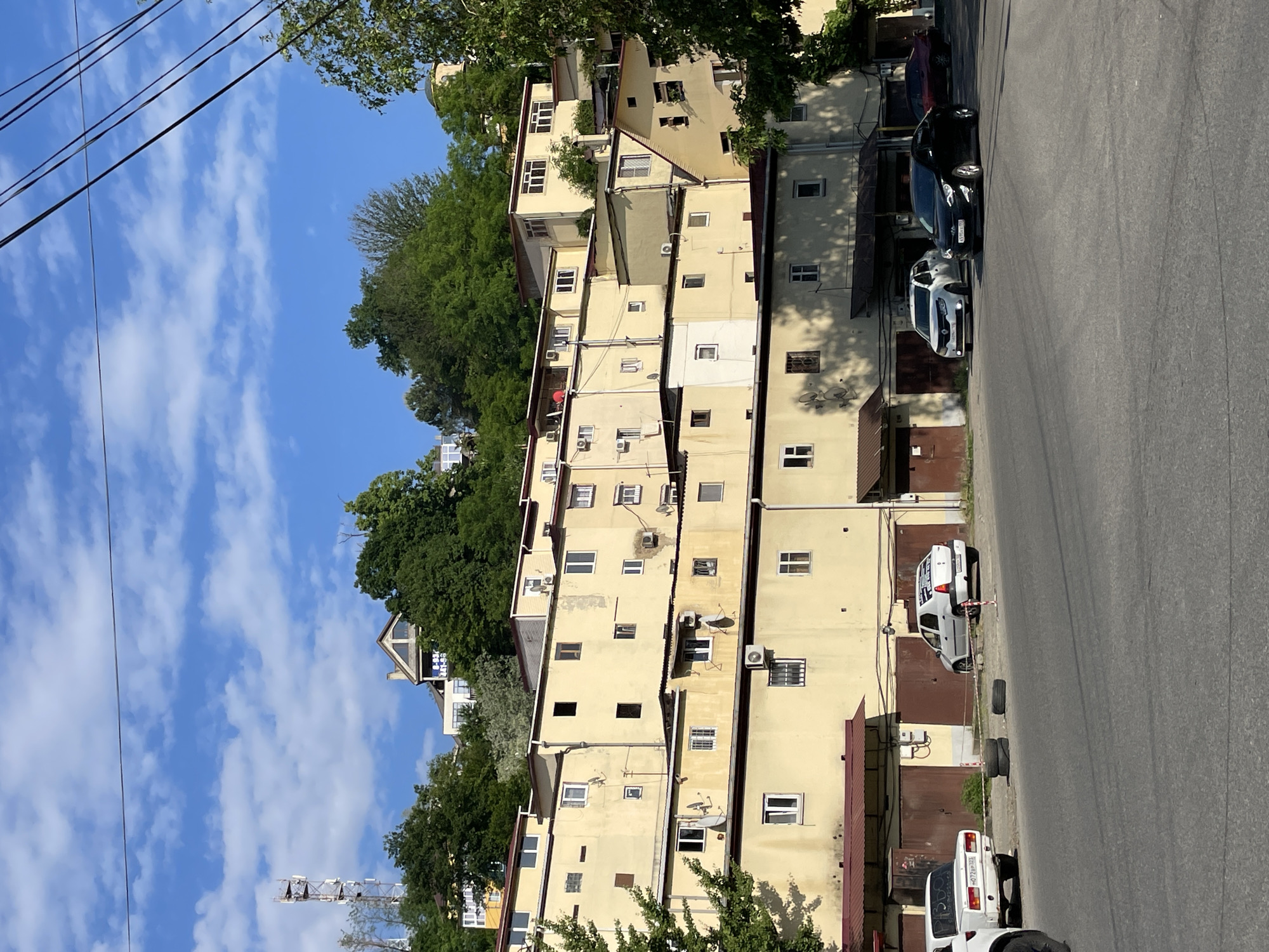 The image depicts a multi-tiered residential complex built into a steep, tree-covered hillside under a clear blue sky with scattered white clouds. The scene is viewed from an asphalt street.
In the foreground, an asphalt road extends across the frame. On the left, a white Lada sedan, license plate `н072вр 123`, is parked facing away from the viewer; its rear window displays text including "Разбор" and a phone number. Three stacked black tires rest on the dirt shoulder next to the Lada. To the right, a white Renault sedan, license plate `н285нр 123`, is parked facing right, with advertisements for "ГРУЗОТАКСИ" and phone numbers on its rear window. A black Hyundai Elantra, license plate `к539хх 93`, is parked angled towards the viewer. A dark red sedan is partially visible behind the Hyundai. Overhead, several black utility lines traverse the sky. No individuals are visible.
The midground is dominated by numerous pale yellow (cream-colored) buildings, stacked in terraces up the slope. These structures feature dark reddish-brown corrugated metal roofs and multiple windows, many with white frames. Several external air conditioning units and satellite dishes are affixed to the building exteriors. Some wall sections exhibit staining and minor paint degradation. At street level, multiple large, brown garage doors are integrated into the ground floor of the buildings.
The background consists of a densely forested hillside. On the upper left, a tall, metal lattice communications tower is visible above the tree line. Further to the right on the hilltop, a building with a prominent gable roof is partially visible, featuring a blue sign displaying a Russian phone number beginning with "8-938-". On the far right, a building with a dome-like roof is partially obscured by trees.
The combination of Russian vehicle license plates (Krasnodar Krai codes 123 and 93), hillside architecture characteristic of coastal cities, and the specific phone number prefix strongly indicates the location as Sochi, Russia. The time of day appears to be daylight hours, with sun creating distinct shadows.