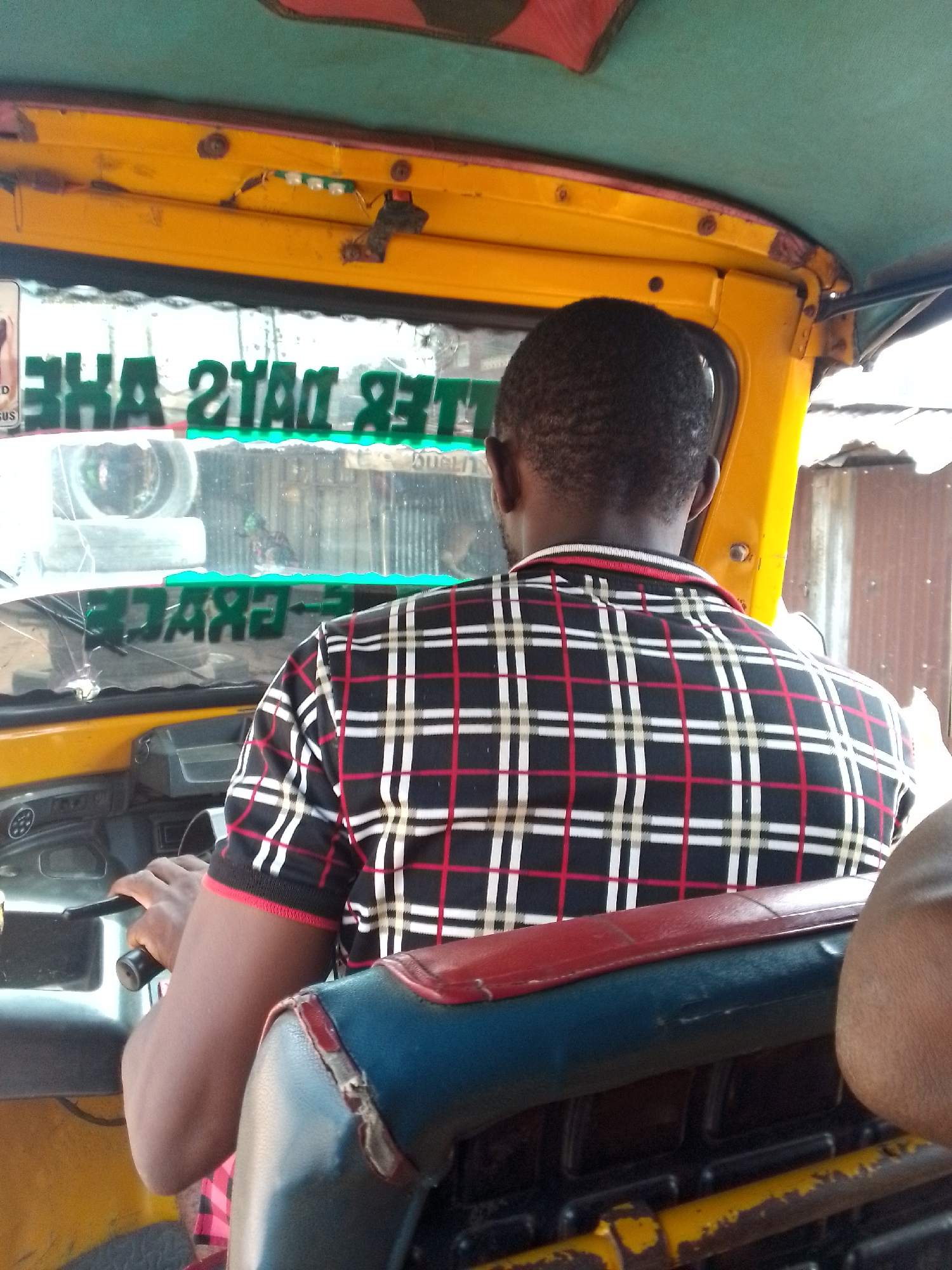 The image shows the interior of a yellow auto-rickshaw, likely in Nkpor, Nigeria. A man, seen from behind, is driving the vehicle. He is wearing a black, red, and white checkered shirt. The auto-rickshaw's dashboard and steering wheel are visible. Green lettering is affixed to the windshield, reading "BETTER DAYS ARE COMING" and "FRACTURES." The interior of the auto-rickshaw is somewhat worn, with some peeling paint and exposed wires visible on the yellow structure above the windshield. The outside environment suggests a somewhat informal urban setting with corrugated metal structures visible.
