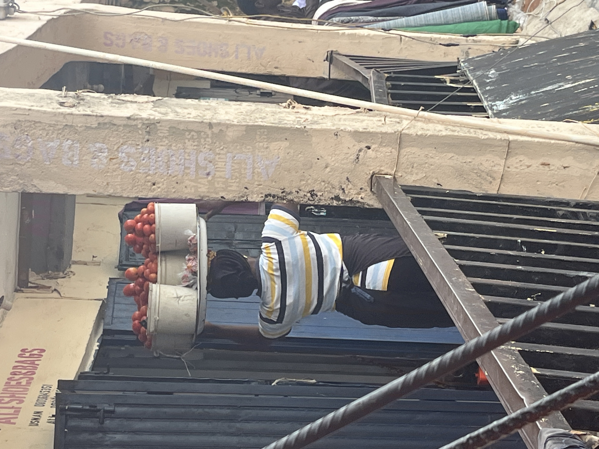 The image depicts a street scene in Jos, Nigeria, focusing on a person standing in front of a shop. The person is wearing a horizontally striped t-shirt with yellow and white stripes and a dark shirt underneath, and dark pants. They are standing next to several stacked white buckets filled with red tomatoes.
Behind the person and the tomatoes, there's a building with multiple levels. The lower level features a shop with a dark, corrugated metal shutter that is partially open. Above this shutter, a section of the building's facade is visible, painted in a light color, with the text "ALI SHOES & BAGS" written in pink lettering, along with a phone number. Further up, there are more architectural elements and what appear to be stacked textiles or fabrics in various colors, suggesting a market or retail area. The overall impression is of a busy commercial environment, potentially a street market. The lighting suggests it is daytime, but no specific time can be determined. The weather appears to be neutral.