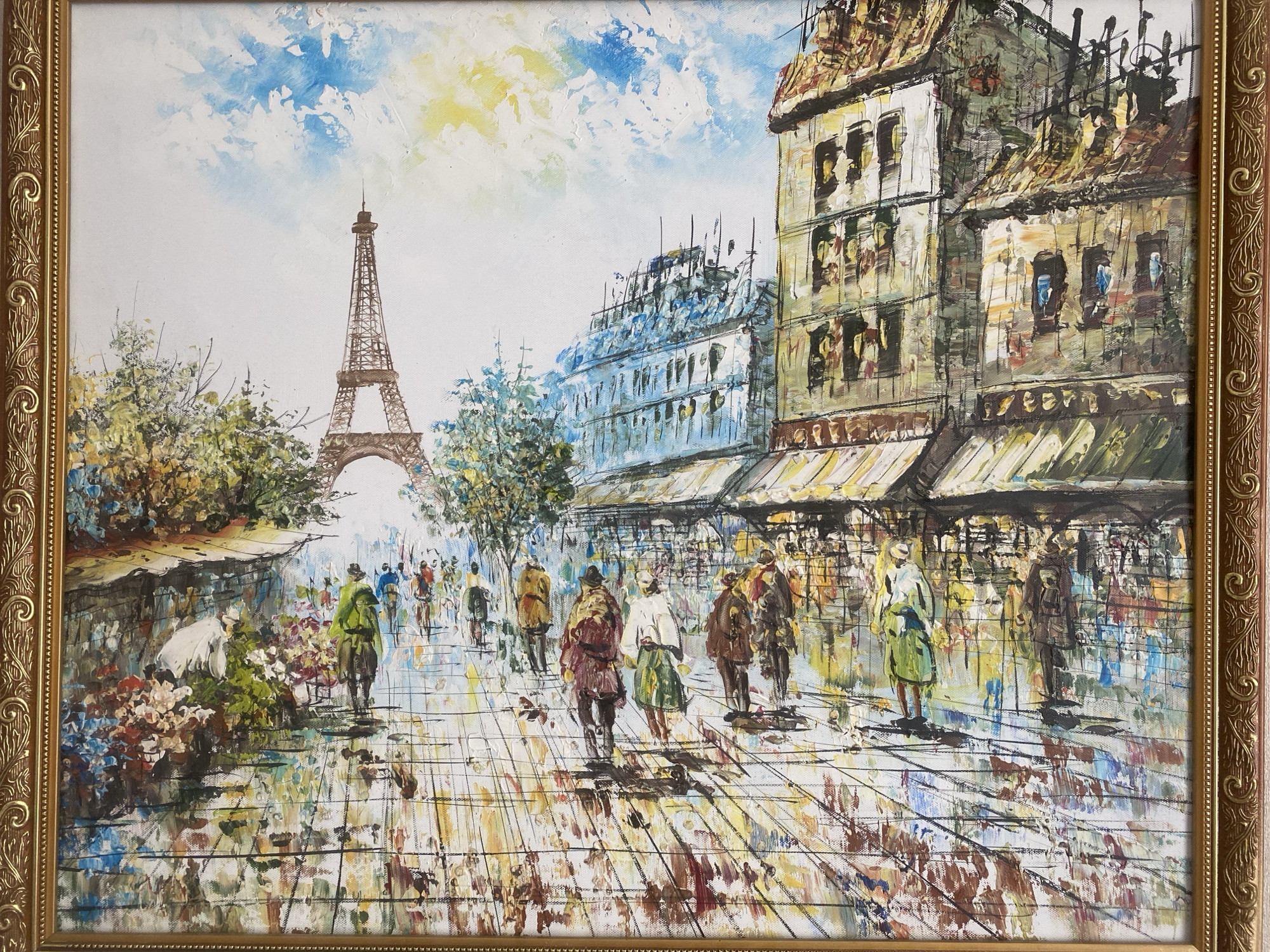 The image is an oil painting of a street scene in Paris, France, featuring the Eiffel Tower in the background. The painting depicts a wet street with reflections, pedestrians walking along the sidewalk, and buildings with awnings and windows. A flower stall is on the left side of the painting. The sky is mostly white with patches of blue and yellow. The painting is framed in a gold-colored ornate frame.