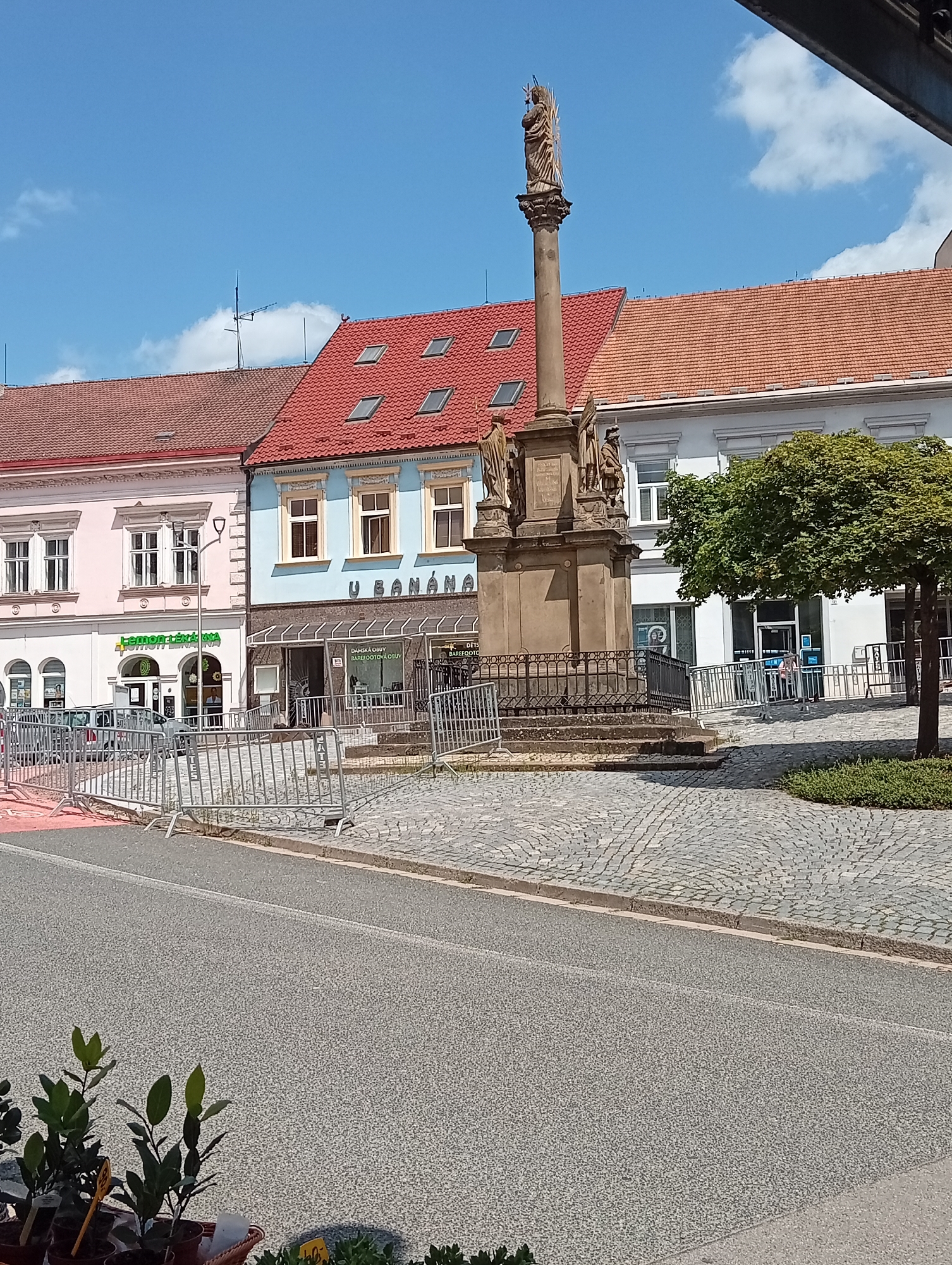 The image depicts a street scene in Tišnov, Czechia, focusing on a cobblestone plaza with a prominent monument. The monument, a stone column topped with a statue, stands in the center of the plaza, surrounded by a black metal fence. Several buildings line the plaza, displaying various architectural styles and colors, with storefronts visible on the ground floor. One building is painted light pink, another sky blue with yellow trim, and a third is white. A large green tree is situated to the right of the monument. Temporary metal barricades are present in the foreground, suggesting a temporary event or construction. The sky is clear blue with a few white clouds. The foreground also shows potted plants.