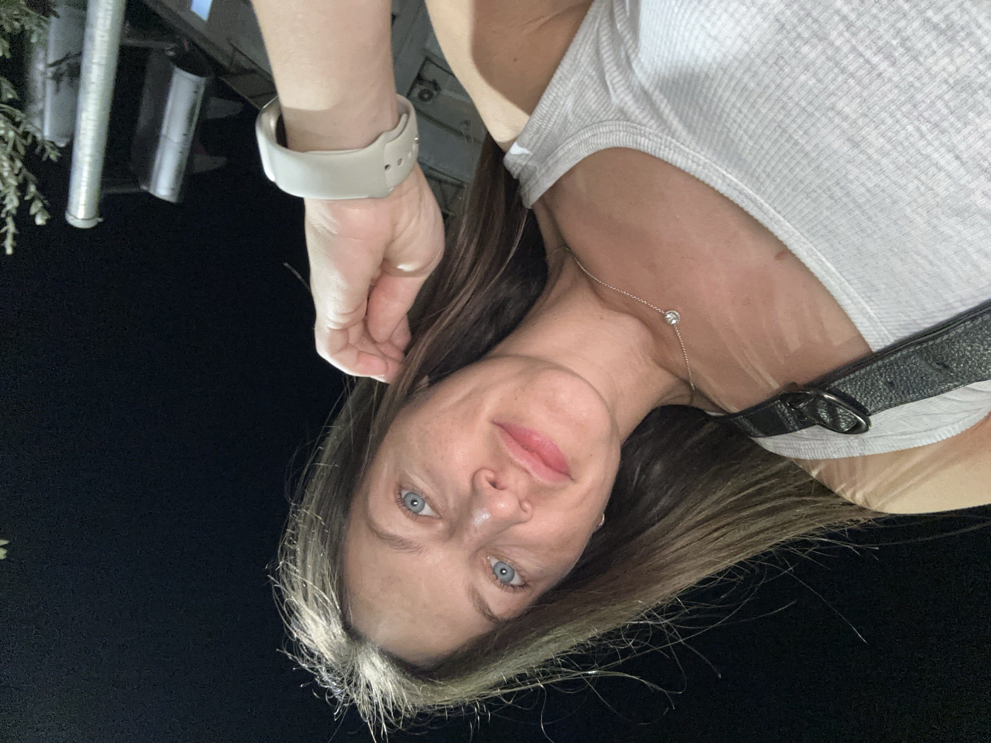 A low-angle, medium shot captures a woman with blue eyes and brown hair looking directly at the camera. She is wearing a white ribbed tank top and a silver necklace with a small pendant. Her left hand is raised, touching her hair. A person's wrist with a light-colored smartwatch is visible in the upper right corner of the frame. In the background, there are out-of-focus elements of what appear to be streetlights or traffic signals. The scene is dimly lit, suggesting it is nighttime, and the environment appears to be outdoors, possibly in an urban or suburban area like Donji Pobori, Montenegro.