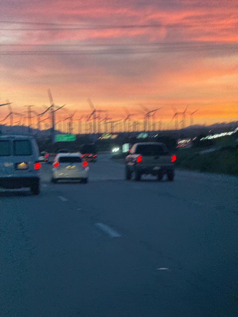This image captures a highway scene at dusk or dawn in the Palm Springs area of the United States. The sky dominates the upper portion of the frame, painted in vibrant hues of orange, pink, and soft purple, indicating either a sunset or sunrise. Several dark power lines stretch horizontally across the upper third of the sky.
In the mid-ground, a vast wind farm is visible on distant hills and mountains, featuring numerous wind turbines with their blades silhouetted against the colorful sky. The turbines appear in various states of motion blur, suggesting they are spinning. The landscape beyond the highway appears arid and mountainous.
The lower half of the image shows a multi-lane highway, where several vehicles are traveling. The scene is somewhat blurry, especially the moving cars, implying the photo was taken from a moving vehicle. In the foreground on the left, the rear of a light-colored van is partially visible. Further down the road in the center, a light-colored sedan with illuminated red taillights is seen. To its right, a dark-colored pickup truck also displays red taillights. White lane markings are faintly visible on the dark asphalt of the road.
The overall impression is one of movement and travel through a landscape characterized by both natural beauty (the sunset/sunrise and mountains) and modern energy infrastructure (the wind farm). No explicit text is clearly visible in the image.