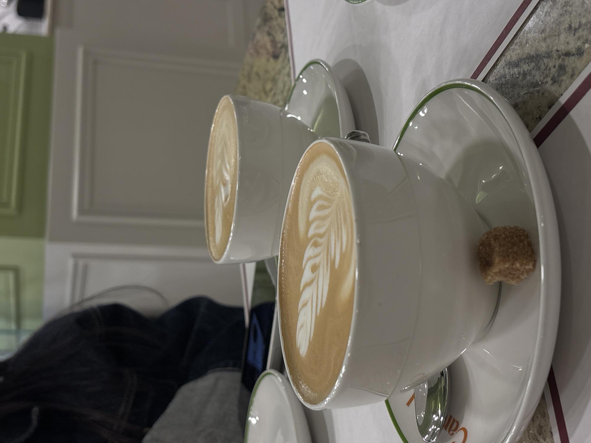 The image shows two cups of cappuccino coffee served in white cups and saucers with a green rim. One cup has a brown sugar cube on the saucer. The coffee in both cups has latte art. The cups are on a white table surface with a gray and white textured edge. A person wearing a dark denim jacket with a gray shirt underneath is partially visible in the lower left of the image. The background shows a light green wall and a white wall with paneling. The location is likely a cafe or restaurant in Yerevan, Armenia based on the appearance of the items and setting.