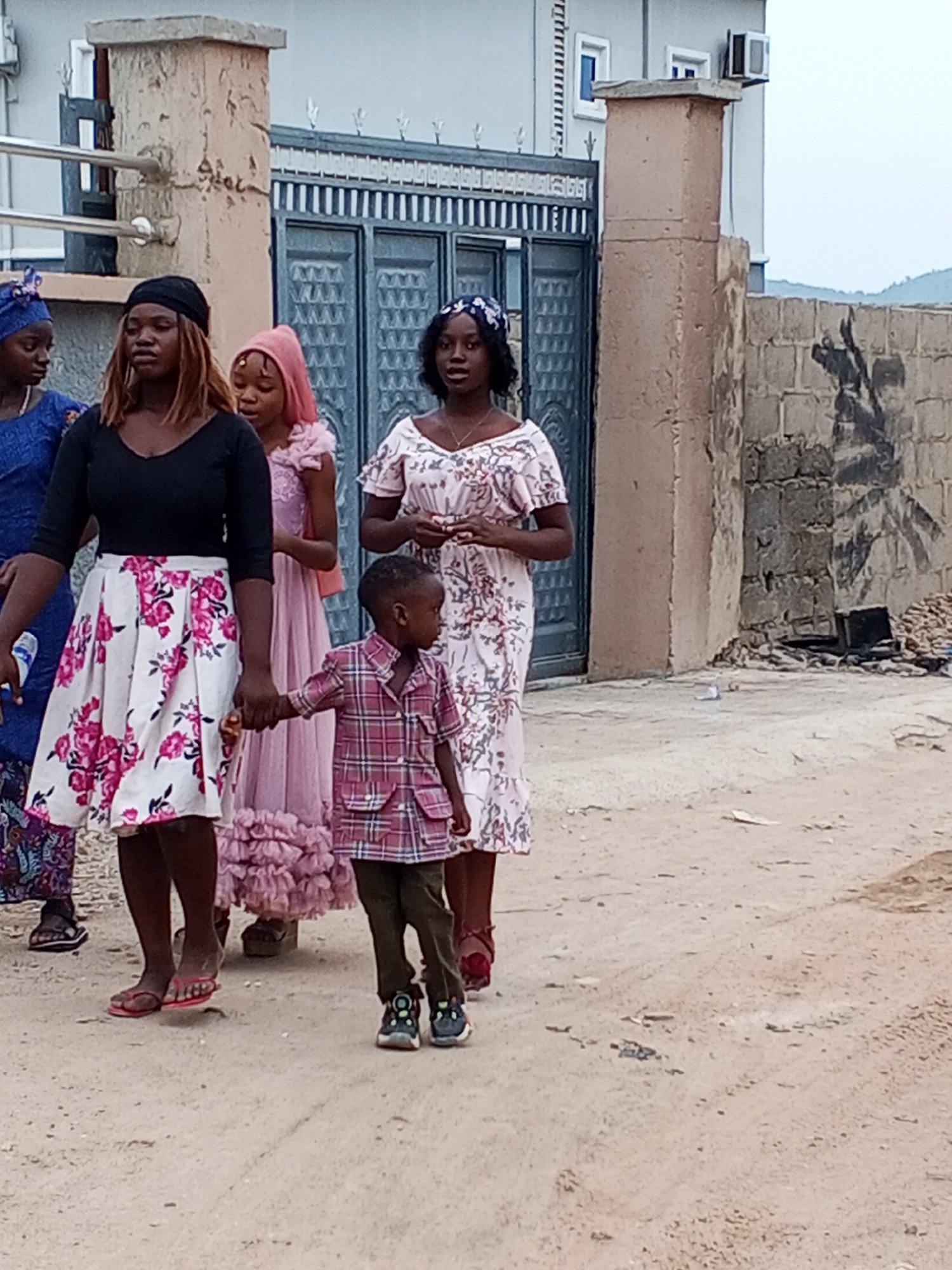 A group of people are walking along a dirt road in Otokiti, Nigeria. The group consists of five women and one young boy. The women are wearing dresses and head coverings, while the boy is wearing a plaid shirt and pants. The woman in the front is wearing a black shirt and a white skirt with pink flowers. She is holding the boy's hand. A building with a blue gate and a block wall are in the background. Some garbage and debris is lying near the block wall.