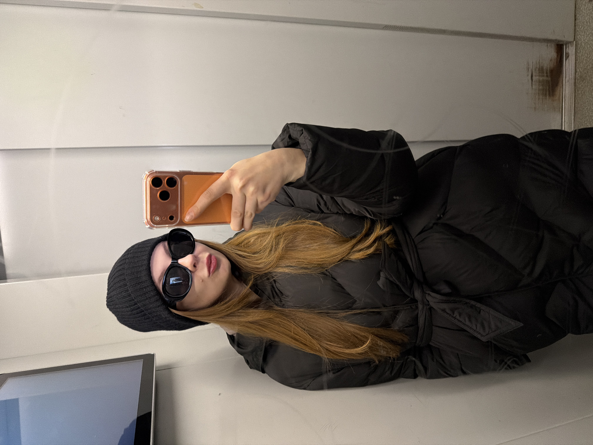 This indoor image from Khimki, Russia, shows a young woman taking a selfie in a reflective surface, likely a mirror. She is positioned slightly off-center and appears to be standing or sitting upright, though the image itself is rotated 90 degrees.
The woman is dressed in a black, belted puffer coat and a black knitted beanie. She wears large, black oval-shaped sunglasses that obscure her eyes. Her long, light brown hair flows over her shoulders and down her back. In her right hand, she holds a smartphone with an orange and clear case, pointing it towards her reflection. Her lips are slightly parted, displaying a neutral or subtly posed expression.
The background consists of plain, light-colored (white or light grey) wall panels with a horizontal seam running through the middle. Some minor scuff marks and discoloration are visible on the panels, particularly in the top right and far right. A portion of a dark digital screen or monitor is visible in the bottom left corner of the frame. The time of day is not discernible from the indoor lighting, but her warm attire suggests cooler weather. There is no visible text in the image.