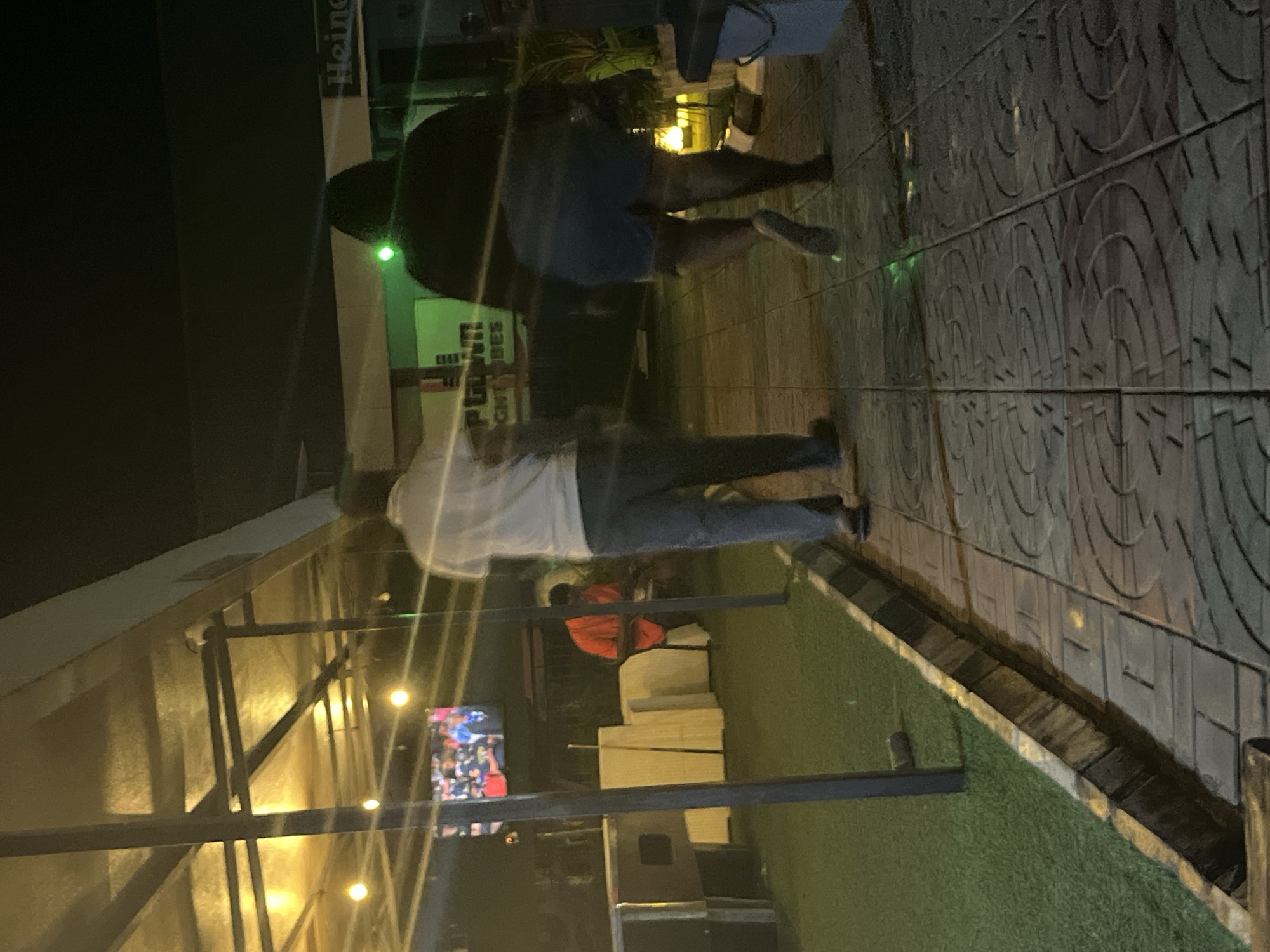 An outdoor scene at night. Two individuals are walking away from the camera on a paved walkway. The person in front is wearing a dark hooded top and blue shorts, and the person behind them is wearing a white shirt and blue pants. In the background, to the left, a man in an orange shirt is seated. A television screen is visible above and to the left of the seated man. To the right, a sign with the word "Heine" is visible. The ground is covered with artificial turf to the left of the walkway and pavers on the walkway. There are overhead lights and some lighting from within structures.