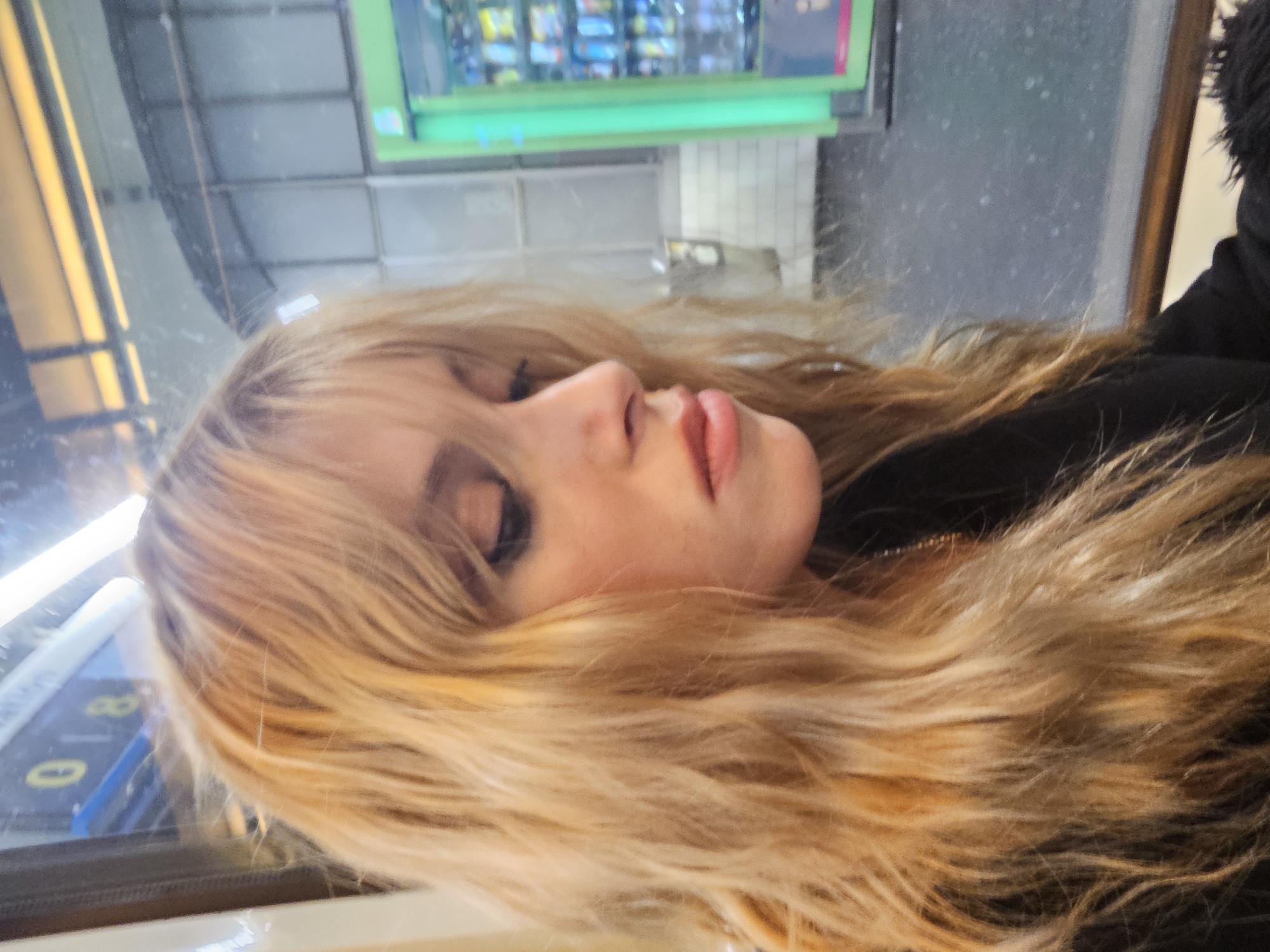 A young woman with long, blonde hair is lying on a bus seat, likely in Paris, France, given the visible vending machine and bus seat design typical of Parisian public transportation. Her eyes are closed, and she appears to be resting. The background shows a blurred reflection of the outside environment, possibly a subway or bus station, with vending machines and white tiled walls.