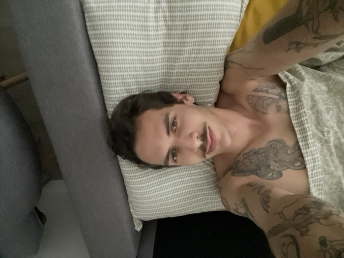 A fair-skinned man with dark hair and a mustache is lying on a striped pillow. He is partially covered by a blanket and has tattoos on his upper body and arms. The background includes a grey couch and a yellow pillow. The photograph is taken from an overhead angle.
