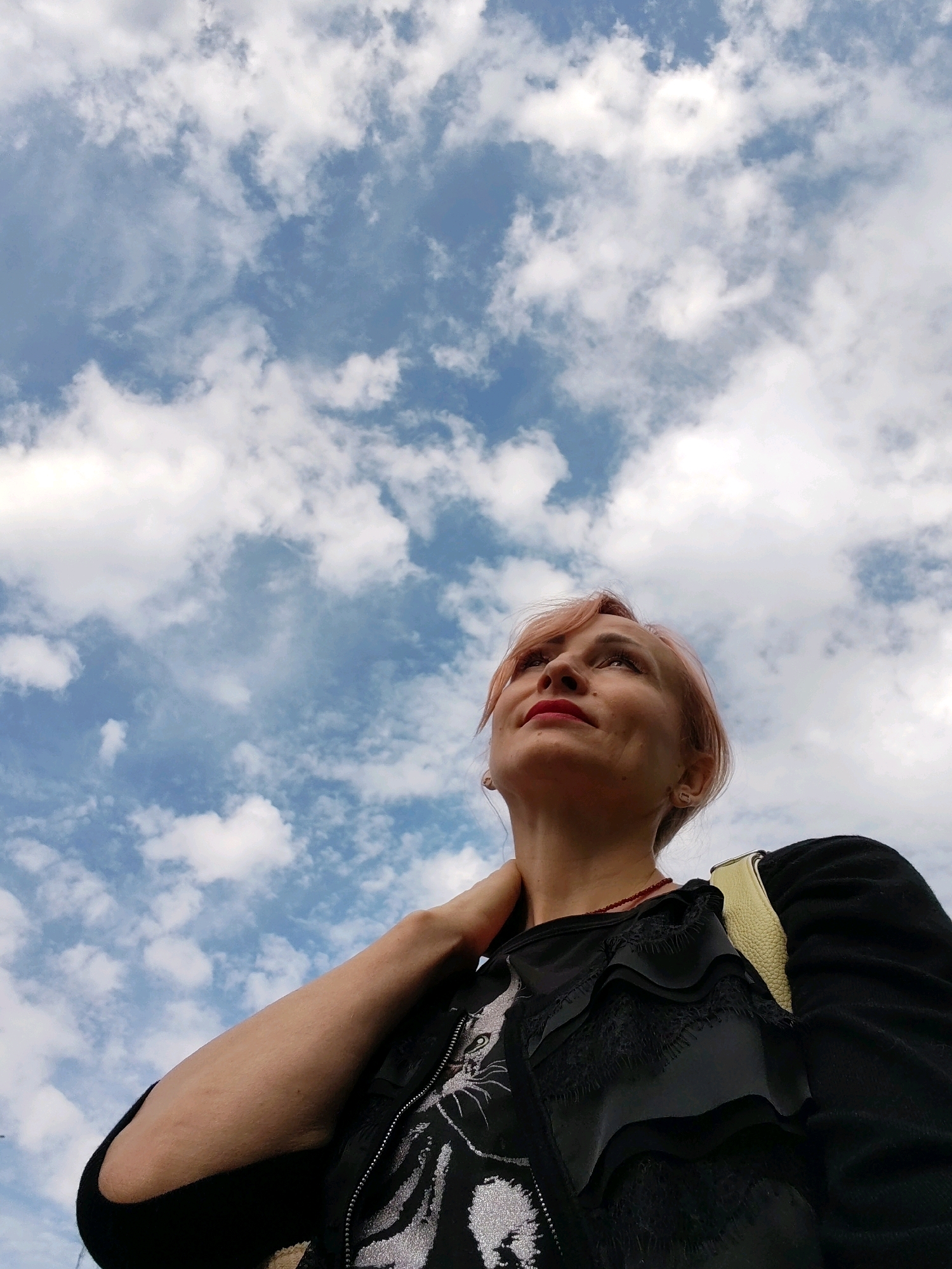 A low-angle shot captures a woman with light pink hair looking upwards against a cloudy blue sky. She is wearing a black top with intricate ruffled detailing on the chest and a silver design. A yellow strap is visible over her right shoulder. Her left arm is bent at the elbow, with her hand resting on her neck. The image appears to be taken outdoors in an urban environment, possibly in the city of Chaikovsky, Russia, given the context. The sky is filled with scattered white clouds.
