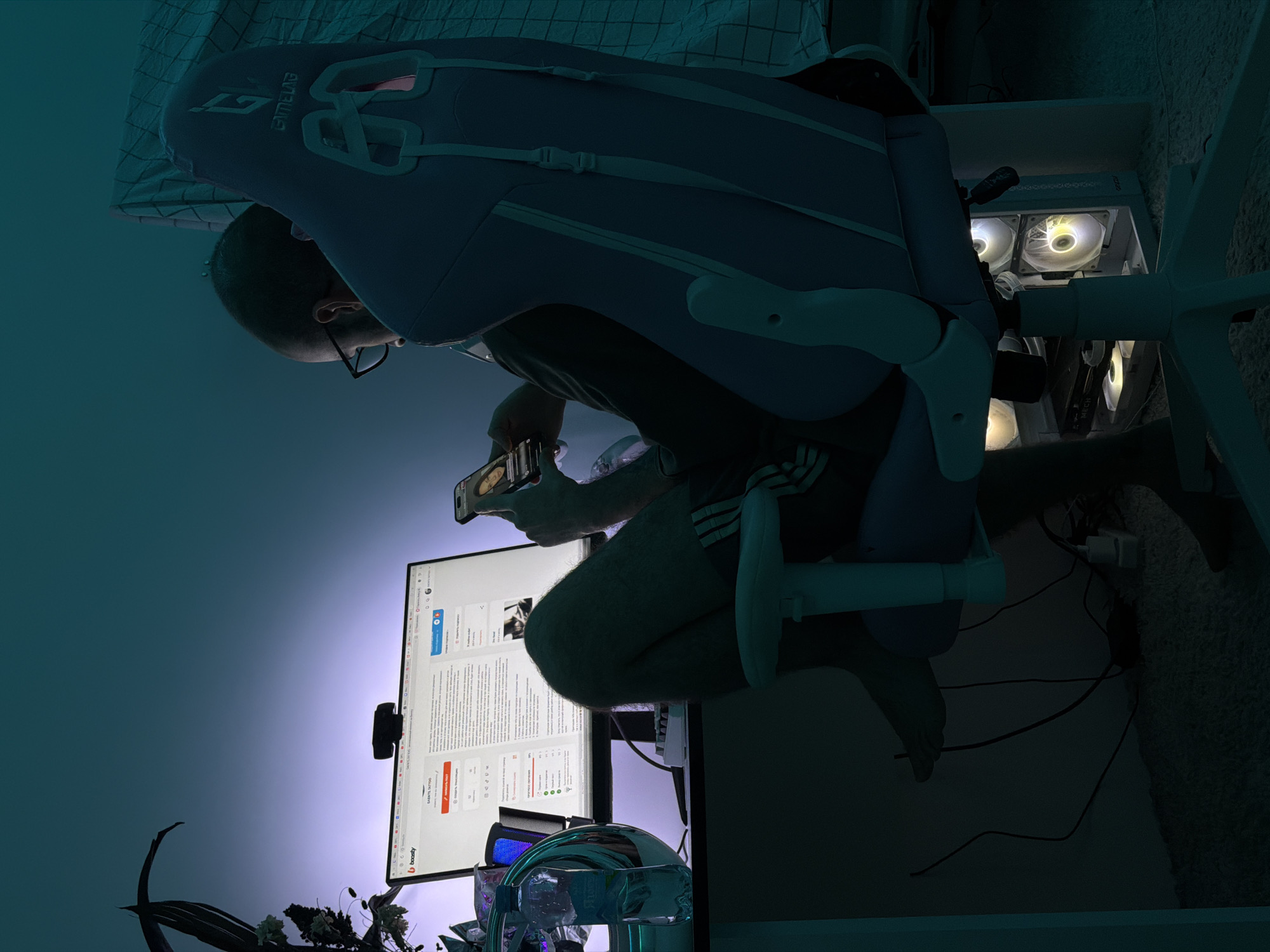 The media file captures an indoor scene, likely a home office or gaming setup, bathed in cool, artificial light during what appears to be the evening or night. The image is vertically oriented but depicts a person seated in a gaming chair at a desk, looking at a smartphone.
The main subject is a person, identifiable as male by arm hair and wearing glasses, seen from behind and to the side. They are sitting in a light blue and white gaming chair with the "GT RACING" logo visible on the headrest, along with "GAME LAB" branding. In their hands, they hold a smartphone, which is illuminated by its screen. Their legs are visible, suggesting they are wearing shorts or athletic wear with striped detailing (possibly Adidas).
In front of the person is a large computer monitor displaying a webpage. The webpage features the "Boosty" logo at the bottom left and a visible title or username "SABTLE TATTOO." The content includes text, images (some cropped out), and UI elements like likes, comments, and shares, with some visible Cyrillic characters. A webcam is mounted on top of the monitor. To the right of the monitor, a white desktop computer tower is visible, with several circular fans inside glowing with yellow/white light, and "MIER" text partially visible on one fan.
The ambient lighting in the room is dark, with a blue and purple gradient emanating from the area around the monitor, creating a moody atmosphere. On the desk, near the monitor, a clear plastic water bottle is present, along with some dark, possibly artificial, foliage or plants. Wires and cables are visible on the light-colored, carpeted floor beneath the desk.
The scene depicts a moment of leisure or work, with the individual engaging with digital content on both a monitor and a smartphone. The location context is Khimki, Russia.