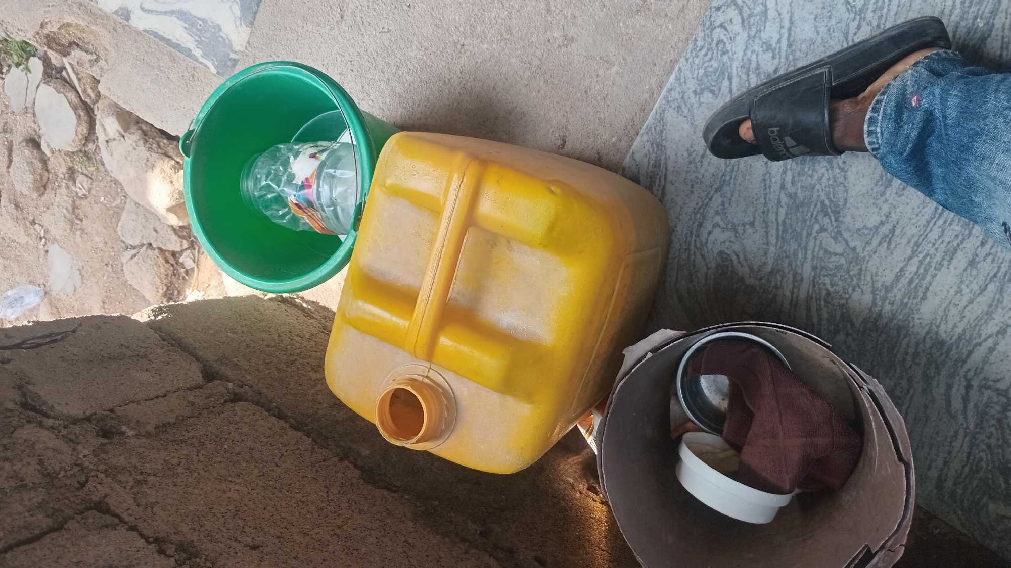 The image displays an outdoor scene in Jalingo, Nigeria, likely during the daytime given the bright lighting and shadows. The setting appears to be an unpaved area, possibly a yard or roadside, with rough, textured ground composed of sand and broken stones.
The central focus is on several objects. A bright yellow plastic jerrycan, commonly used for storing liquids, lies on its side. Next to it, a green plastic bucket contains a partially filled, clear plastic water bottle. To the right, a dark-colored open-top container holds a few items, including what looks like a small metal bowl, a white plastic lid or cup, and a folded piece of brown cloth. A person's foot, wearing a black sandal and blue jeans, is visible in the upper right portion of the frame, suggesting someone is present but out of view.
The objects are arranged in a way that implies they are being stored or are in temporary use in this outdoor location. There are no explicit indications of a specific event, but the arrangement of containers and the presence of a water bottle suggest practical, everyday activities related to water storage or usage. The overall impression is one of a functional, perhaps utilitarian, outdoor space in a Nigerian context. No discernible text is visible on the objects.