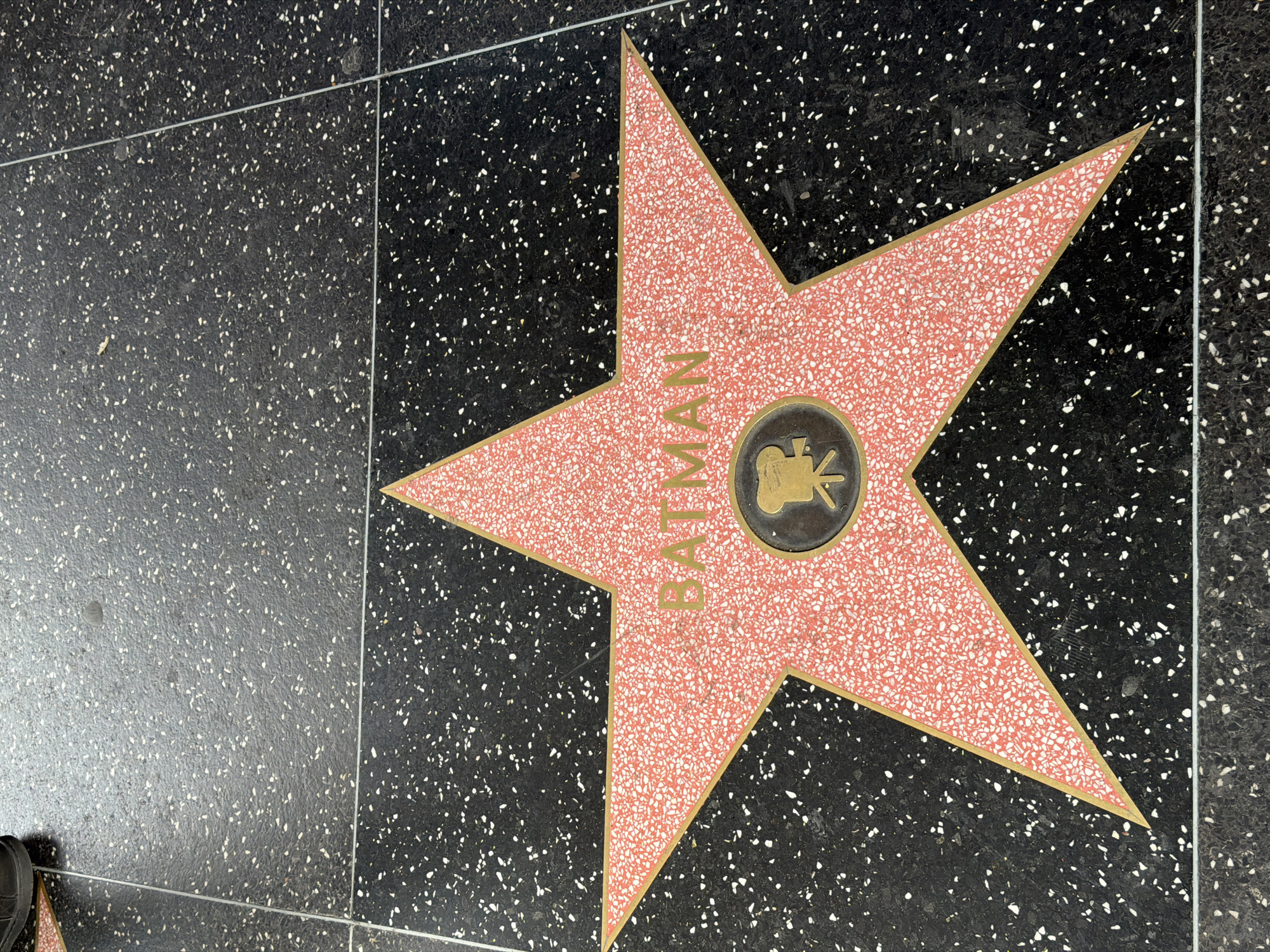 The image shows the star of Batman on the Hollywood Walk of Fame in Los Angeles, California, United States of America. The star is coral pink terrazzo, edged with brass, and embedded in a dark gray terrazzo sidewalk speckled with white aggregate. The name "BATMAN" is inscribed in brass letters within the star. A round brass emblem, depicting a film camera, is at the center of the star. The surrounding sidewalk is divided into squares by lighter gray dividers. A black shoe appears partially in the lower left corner.