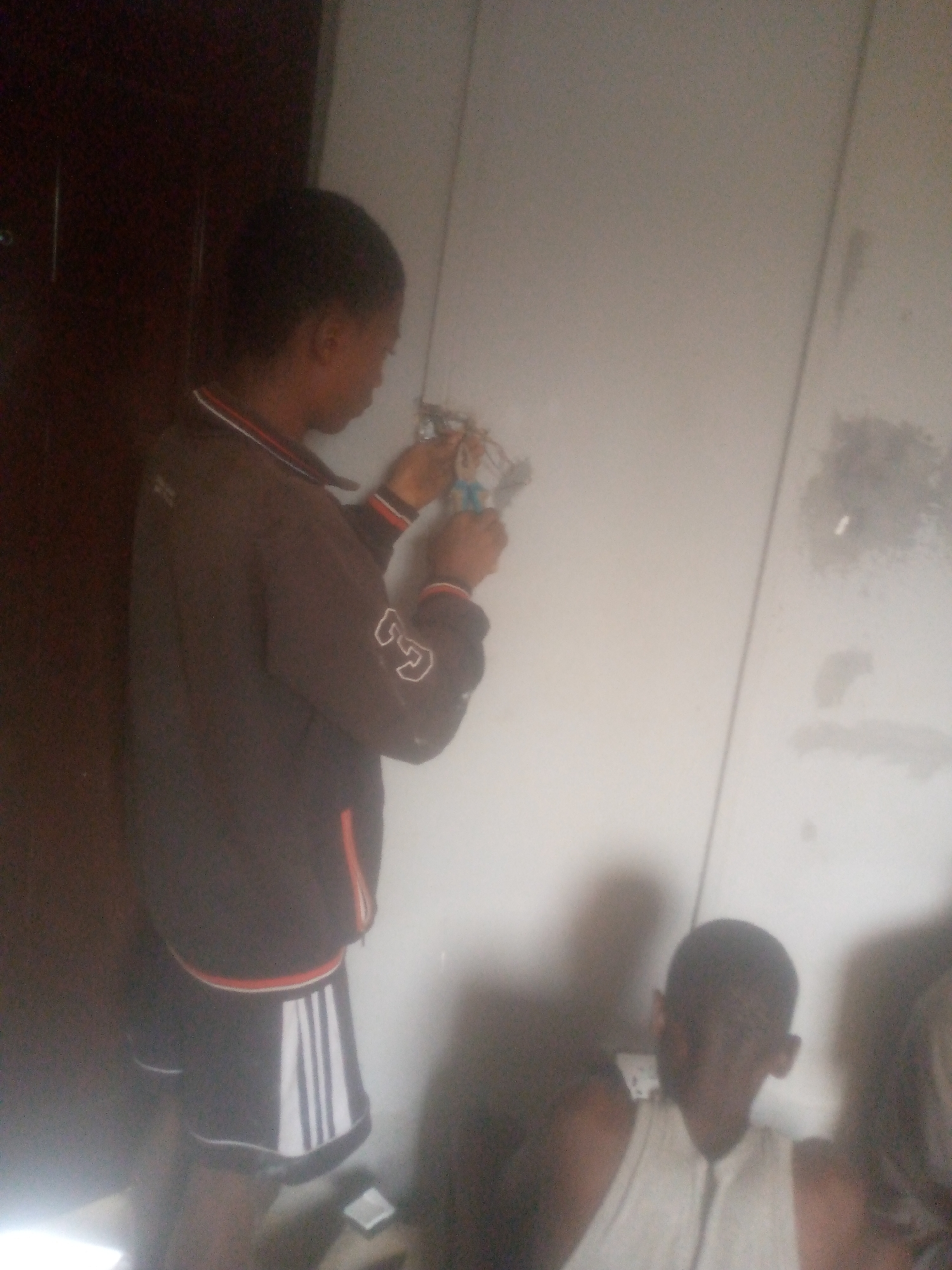 The image displays an indoor scene featuring at least two young individuals, possibly more, engaged in an activity. In the center-left, a young person, likely male, stands facing a light-colored wall. They are actively manipulating what appears to be exposed electrical wires or components on the wall, holding them with their hands. This individual wears a dark long-sleeved jacket or top with red and white striped accents on the collar and cuffs, and a white, illegible graphic on the right sleeve. They also wear dark shorts with distinctive white vertical stripes on the side.
In the bottom right corner of the frame, another young person, also likely male, is partially visible, seated. They wear a light-colored, possibly white, sleeveless top. Their face is somewhat obscured by blur, but they appear to be looking towards the viewer or slightly down. A third person's head is faintly visible further to the right.
The setting is an interior room, with a dark wooden door on the far left and a plain, light-colored wall that shows some smudges and marks, particularly on the right side. The lighting is somewhat dim, suggesting an indoor environment without strong natural light. The primary activity is the handling of electrical elements on the wall, indicating repair, installation, or maintenance work. No discernible text is visible on clothing or in the environment. The image quality is low with some blurriness.