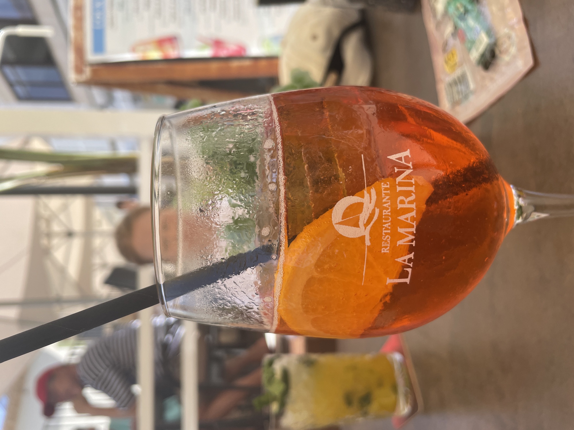 A close-up, eye-level shot focuses on a glass of orange aperitif cocktail with a slice of orange and mint sprigs, garnished with a black straw. The glass is condensation-covered, suggesting it's a cold beverage. In the blurred background, two patrons are seated at a table under a white canopy, characteristic of outdoor dining at a beachside restaurant. The restaurant's logo, "LA MARINA," is visible on the glass. The setting appears to be a restaurant in Santanyí, Spain, possibly near a beach, given the casual attire and outdoor seating.