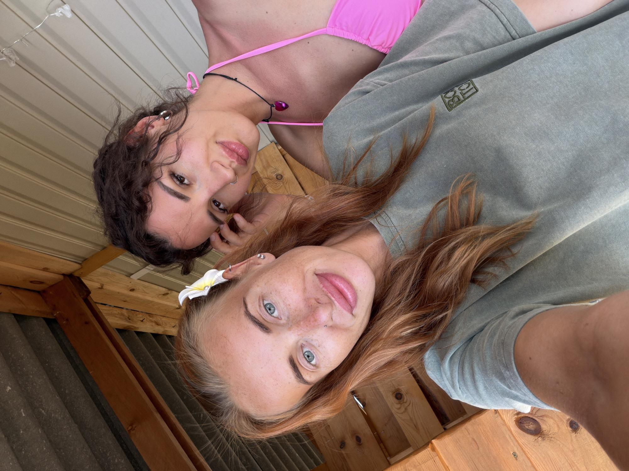 Two young women are taking a selfie outdoors. The woman in the foreground has long, reddish-blonde hair, blue eyes, and freckles. She is wearing a green t-shirt. A white flower is tucked behind her ear. The woman behind her has dark, curly hair and is wearing a bright pink bikini top. She is wearing a black necklace with a pink heart pendant. They appear to be in a rustic, outdoor setting, possibly on a porch or deck, with wooden structures and a corrugated metal roof visible in the background. The overall impression is a casual, summery scene.