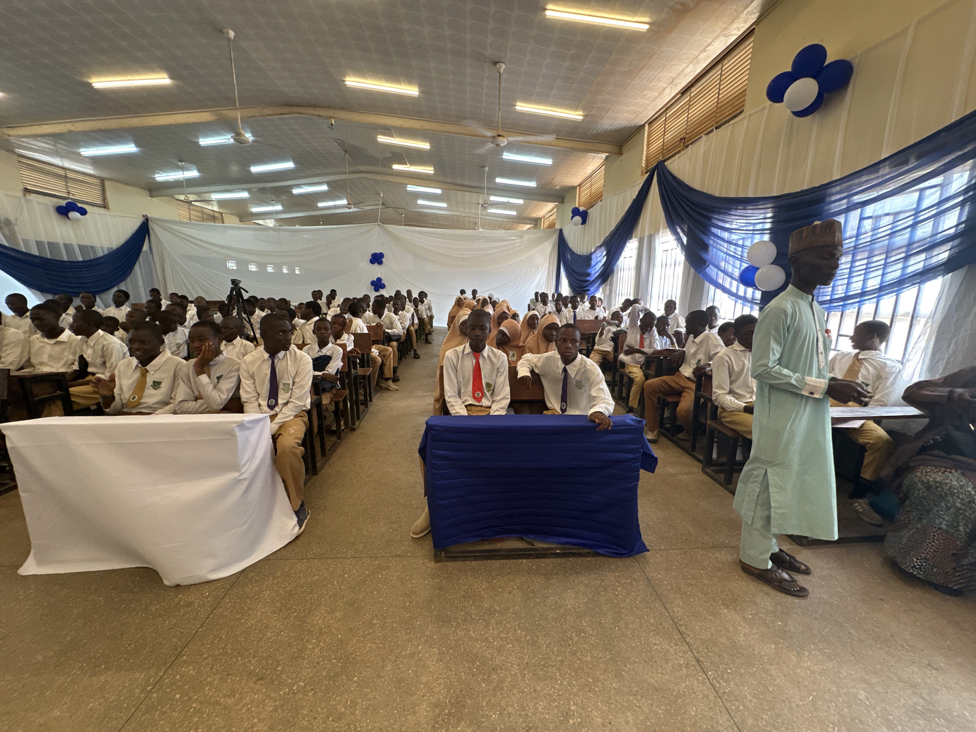 This image depicts an indoor event taking place in a large hall, likely a school or community center, in Unguwan Dosa, Nigeria. The hall is festively decorated with white and blue drapes along the walls and blue and white balloons, suggesting a formal or celebratory occasion.
Numerous young people, primarily students, are seated in rows of wooden desks and benches, attentively facing the front. Most students are wearing white shirts, some with ties in various colors (red, purple, blue) and school crests, along with tan or khaki trousers. A few female students are also visible, some wearing light-colored hijabs. In the foreground, two pairs of students are seated behind low tables covered with white and blue cloths respectively, positioned prominently at the front of the seating area. This setup suggests they might be participants in a quiz, debate, or a similar competition or presentation.
On the right side of the frame, a man, possibly an adult official or teacher, stands facing the audience. He is dressed in a light blue kaftan (agbada) and a matching cap, typical attire in Nigeria.
The hall features a high ceiling equipped with multiple fluorescent light fixtures and ceiling fans. Natural light enters through high windows with louvers, visible above the decorative drapes. A camera on a tripod stands in the background, indicating that the event is being recorded. The floor is covered with light-colored tiles. The general atmosphere appears organized and attentive. While school crests are visible on some uniforms, no legible text is discernible elsewhere in the image. The event is taking place during daylight hours, indicated by the combination of artificial and natural lighting.