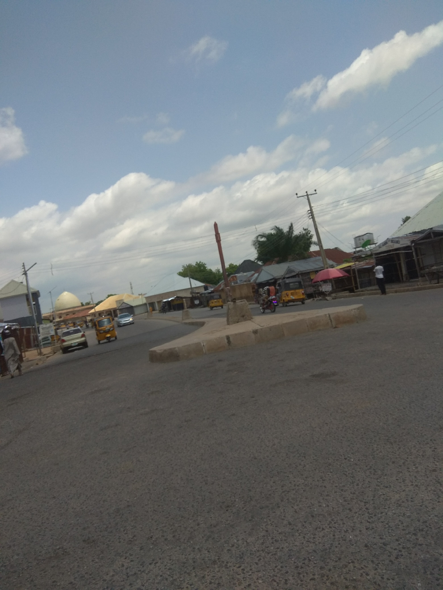 The image depicts a street scene in Jalingo, Nigeria, under a partly cloudy sky. The time of day appears to be daytime, likely late morning or early afternoon, given the brightness and the presence of shadows. The weather is fair, with a mix of blue sky and scattered white clouds.
The foreground is dominated by a paved road, suggesting a relatively urban or semi-urban environment. In the middle ground, a roundabout is visible, a common feature in road intersections. Several vehicles are present, including a yellow auto-rickshaw (also known as a "keke") and a white car. Another auto-rickshaw is seen turning around the roundabout. A few people are also visible, one walking on the sidewalk to the left and another standing near some market stalls on the right.
The background features a collection of buildings, some with corrugated metal roofs, indicative of typical architecture in this region. One building has a distinctive dome shape. There are also signs of commercial activity, with market stalls set up along the street, one with a red umbrella. A utility pole with electrical wires is also visible, a common element in populated areas.
The overall impression is that of a bustling, everyday scene in a Nigerian town. There are no discernible emotions conveyed, as the focus is on the general activity and the environment. No visible text is present in the image.