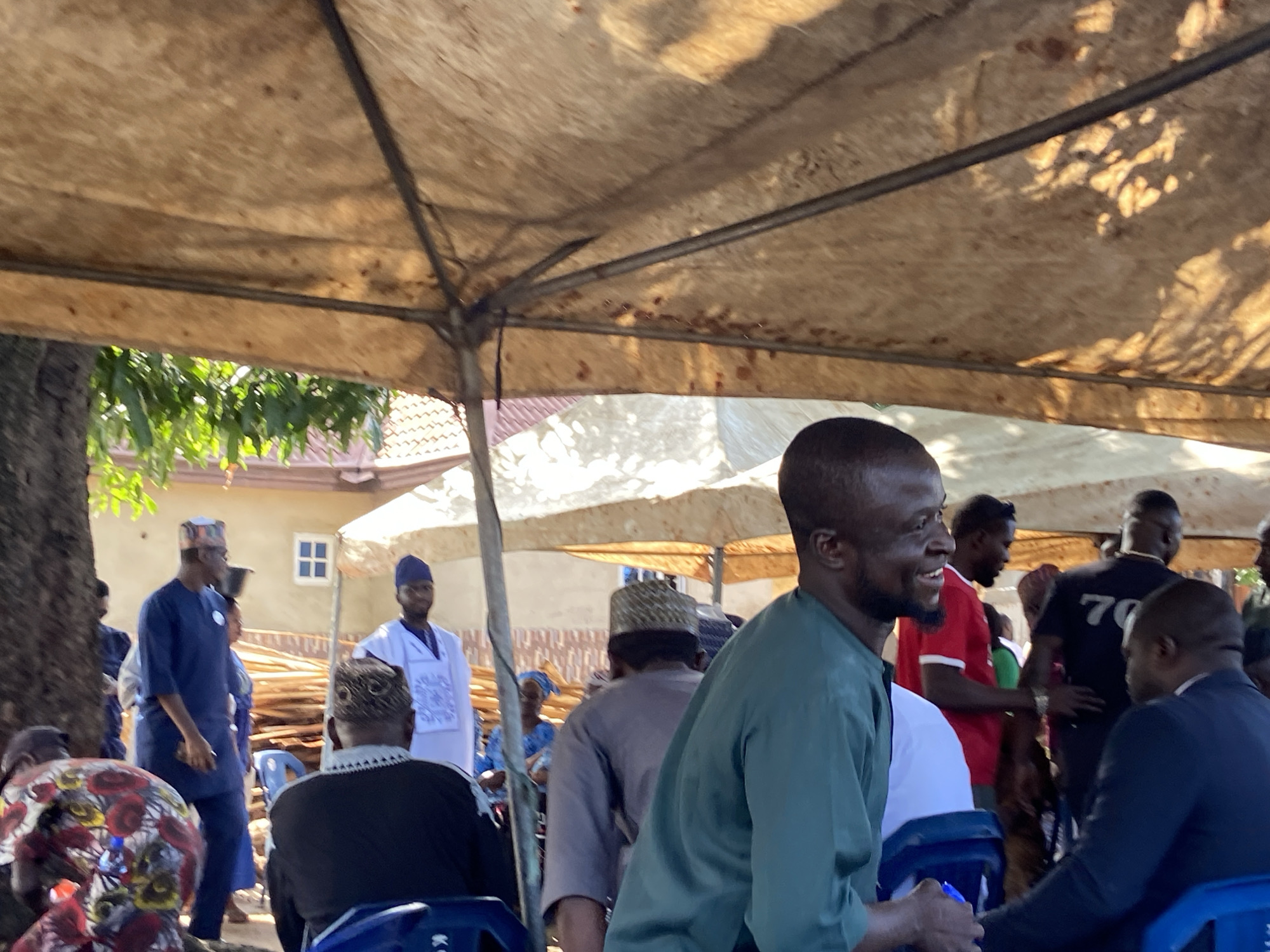 The image depicts an outdoor social gathering under a light-brown canvas canopy, presumably in Mararaba, Nigeria. Bright daylight illuminates the scene, casting shadows through the canopy and from surrounding elements. In the immediate foreground, a Black male in a dark green long-sleeved tunic smiles, looking towards the right, with part of another individual in a blue shirt visible in the lower right, suggesting an interaction.
Multiple individuals are present throughout the mid-ground. To the left, a Black male in a dark blue traditional tunic and cap stands, holding an object resembling a phone. Behind him, a large tree trunk is visible. Further back, a building with a brown tiled roof and a white-framed window is partially seen. To the right of the man in blue, another Black male in a white traditional tunic and a dark blue cap stands. Piles of wooden planks or logs are visible behind him.
Other figures include a Black male in a red t-shirt, standing to the left of the smiling man in green. Behind them, a Black male wearing a black t-shirt with the white number "70" on the back is visible. Several other individuals are partially visible, some seated on blue plastic chairs, and some standing. One seated individual in the lower left wears a colorful patterned head covering and garment. The overall atmosphere suggests a communal event or meeting taking place outdoors under shade.