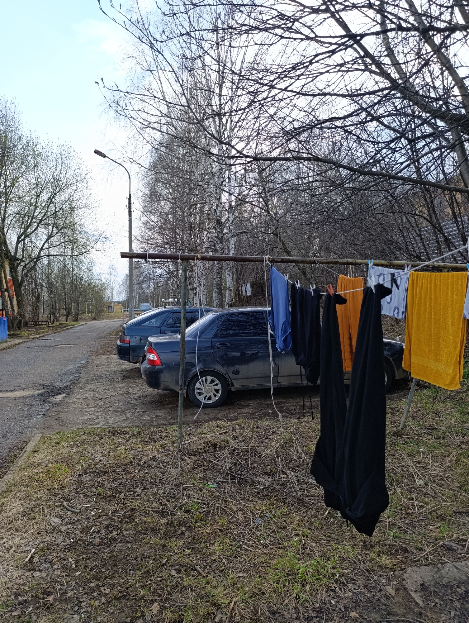 The scene depicts a residential area in Большой Букор, Russia, with a dirt patch alongside a cracked asphalt road. Two dark-colored Lada sedans are parked partially on the dirt, their exteriors appearing grimy. A makeshift clothesline constructed from a metal bar and thin rope stretches across the frame, obscuring parts of the vehicles. Laundry hangs from the line, including blue shirts, black pants, and yellow towels, secured by clothespins. The ground is covered with dry grass, fallen leaves, twigs, and several discarded cigarette butts. Bare trees dominate the background, suggesting early spring or late autumn. A streetlight is visible in the middle distance. The overall impression is one of neglect and modest living conditions.