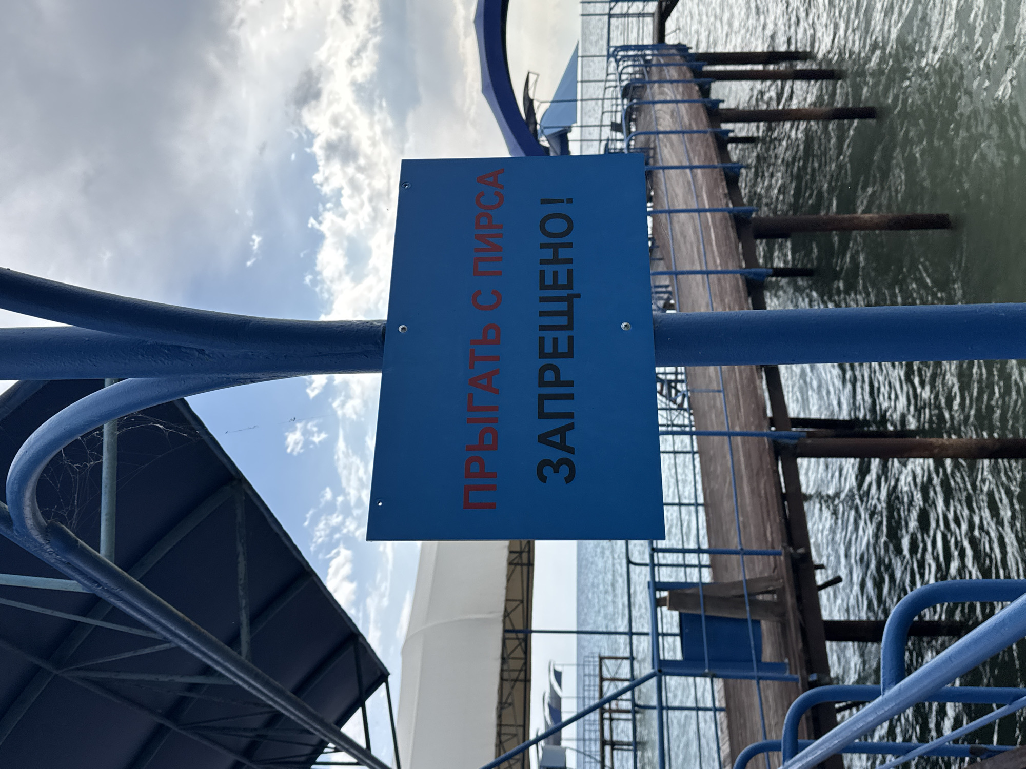 This is a medium shot, eye-level, outdoor, vertical photograph of a blue sign with Russian text attached to a blue metal pole. The sign, positioned centrally in the foreground, has two lines of text. The top line is in red and reads "ПРЫГАТЬ С ПИРСА", and the bottom line is in black and reads "ЗАПРЕЩЕНО!". The sign is attached to a blue pole which is part of a larger blue structure that extends upwards and to the left, appearing to be a covered walkway or canopy. Behind the sign, a wooden pier with blue railings extends into the water. The pier is supported by dark pilings. The water is choppy and greyish-green. In the background, there is a cloudy sky with patches of blue. The overall setting appears to be a waterfront location, likely a pier or a beach facility. The sign indicates that jumping from the pier is prohibited. The location is identified as being in the city of Sary-Oy, Kyrgyzstan.