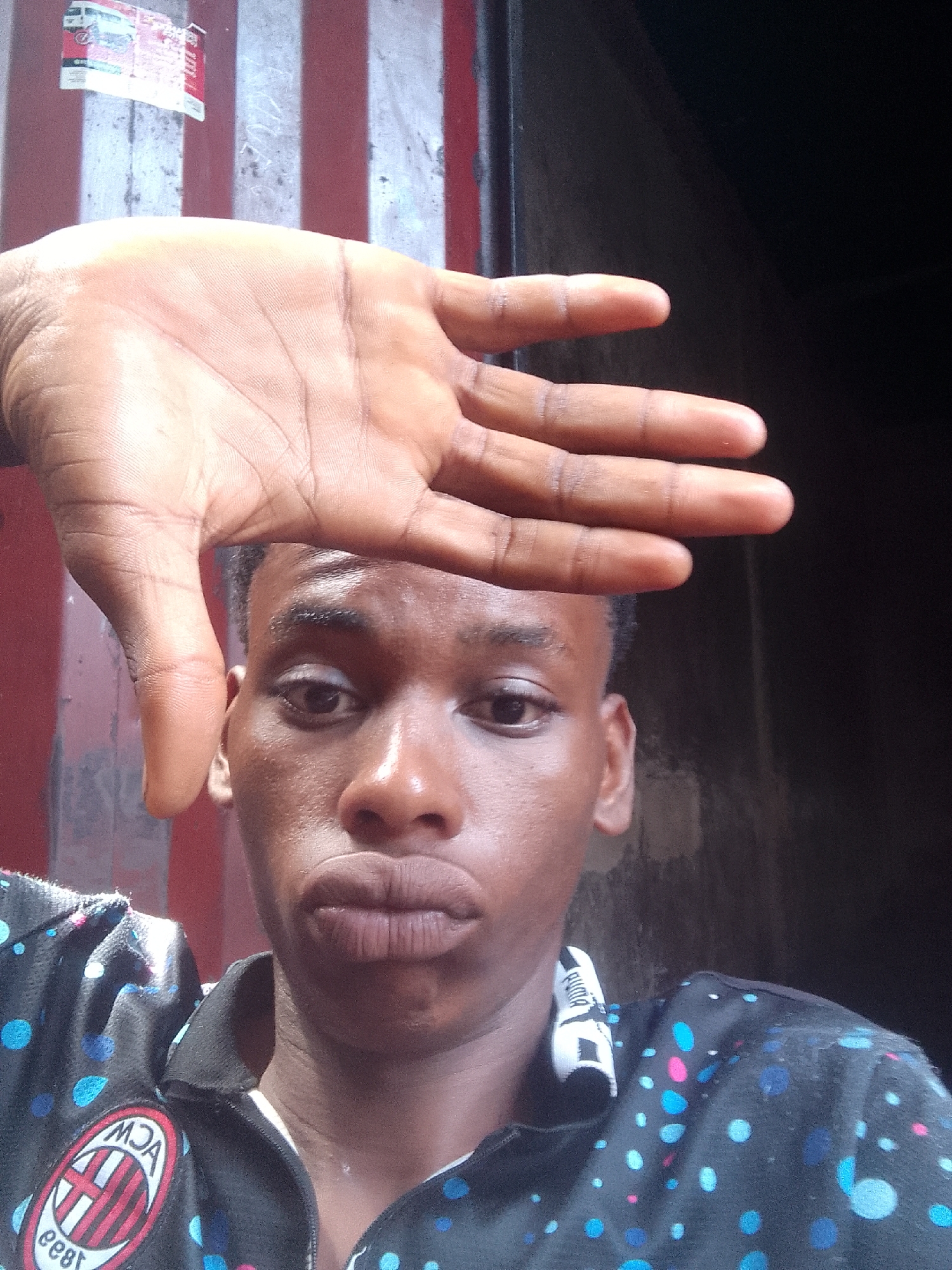 A young man is in a selfie. The man is dark-skinned. The man is wearing a dark polo shirt with light blue and pink polka dots. The man's right hand is in front of his face. The background is a red and white striped wall and dark interior. The image was taken in Onitsha, Nigeria, given the presence of the local advertisement sticker on the wall.
