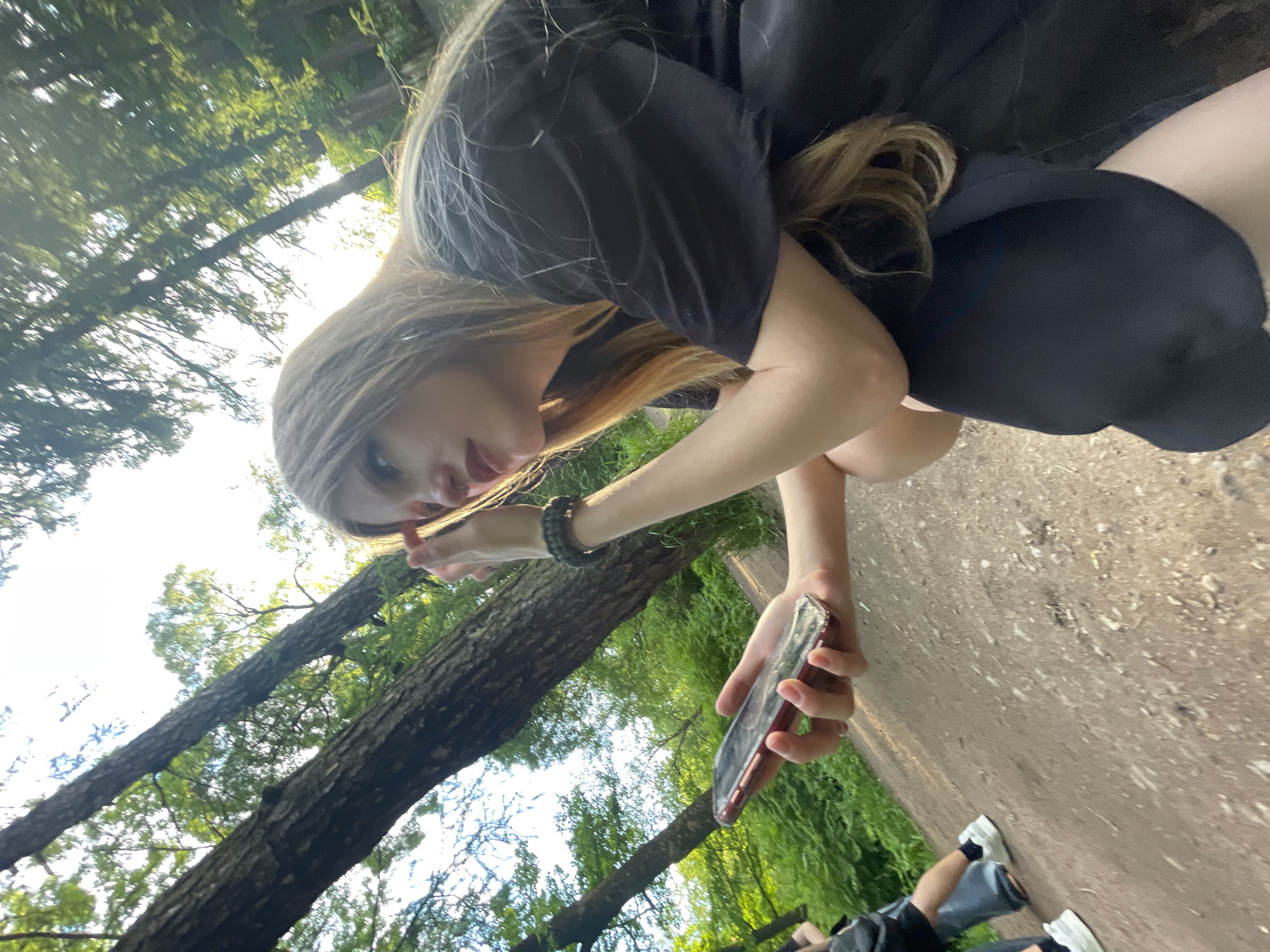This image, taken in what appears to be a park in Saint Petersburg, Russia, captures a casual outdoor scene on a bright, sunny day. The primary subject is a young person, likely female, with long, light brown or blonde hair, seen from a low angle as she leans forward. She is wearing a black t-shirt and dark shorts, with a dark green, possibly paracord, bracelet on her left wrist. Her left hand is raised towards her face, near her mouth, while her right hand firmly holds a smartphone encased in a translucent reddish cover. Her gaze is directed downwards, suggesting she is looking intently at something or someone just out of frame, or perhaps at the camera.
The background features tall trees with abundant green foliage, indicative of a lush natural environment. The ground beneath the subjects is a light brown, possibly dirt or gravel path. In the lower right corner, the legs and feet of another person are visible, wearing dark shorts and white sneakers, suggesting they are seated or resting on the ground nearby. The overall impression is one of relaxed observation or interaction within a park setting during pleasant weather. No visible text is identifiable in the image.